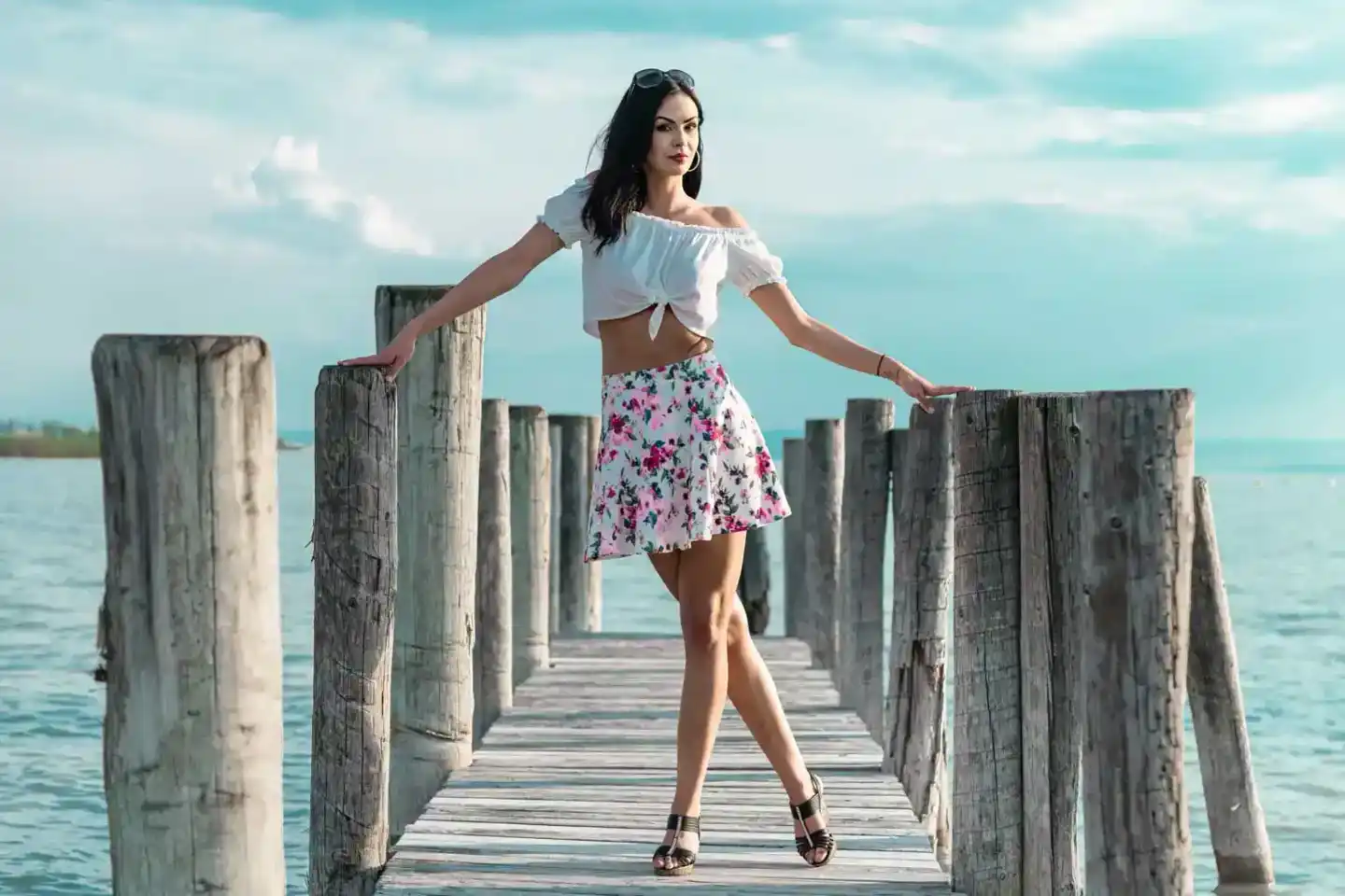 Junge Frau in weißem Crop Top und Blumenrock posiert elegant auf einem Holzsteg am Meer – professionelles Portraitfotografie 