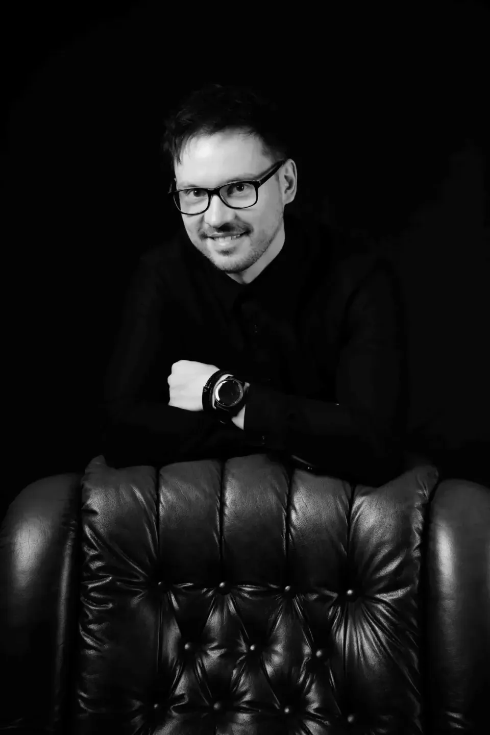Professionelles Portrait eines Mannes mit Brille in schwarzer Kleidung, sitzend auf schwarzem Ledersessel – Portraitfotograf