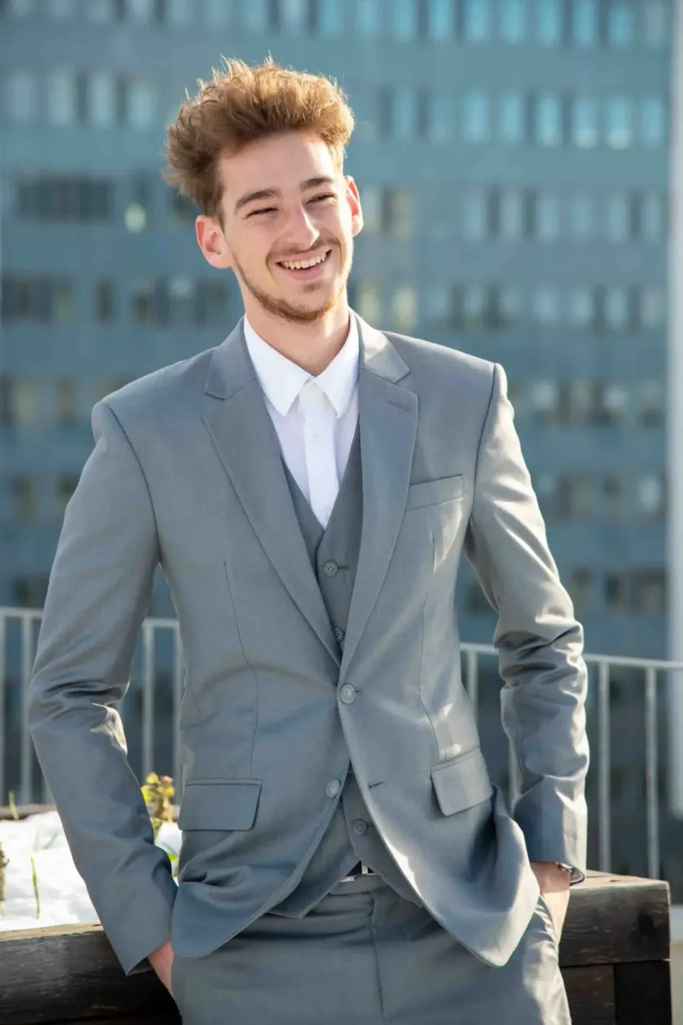Lächelnder junger Mann im hellgrauen Business-Anzug mit weißem Hemd vor modernem Hochhaus – professionelles Headshot für Corp