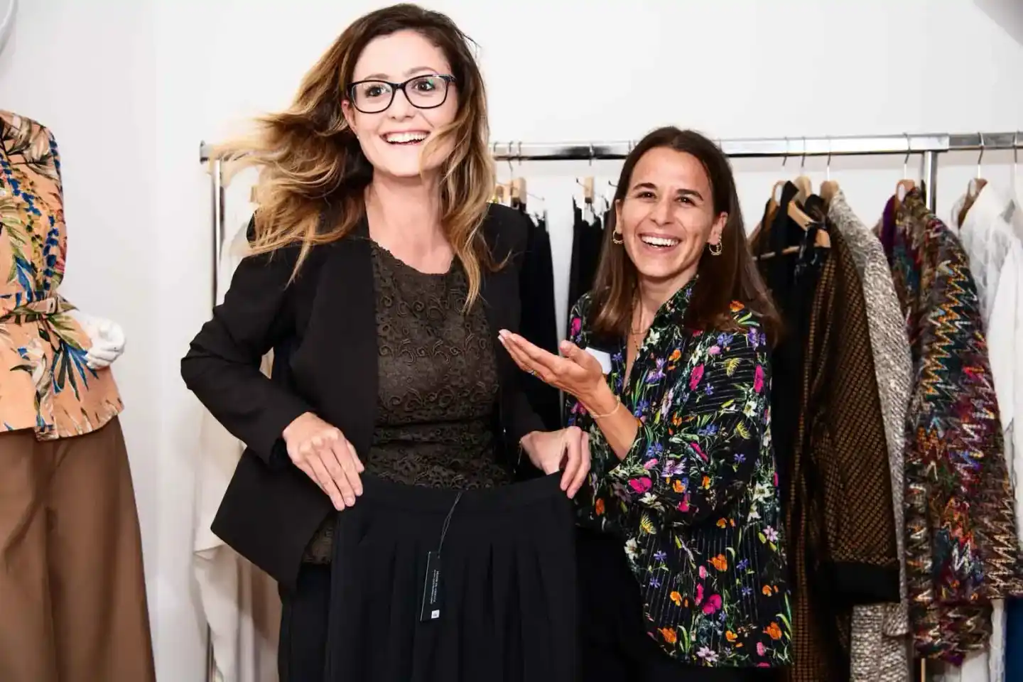 Zwei Frauen lachen vor einer Kleiderstange mit bunten Designs bei einer Modeausstellung in Wien