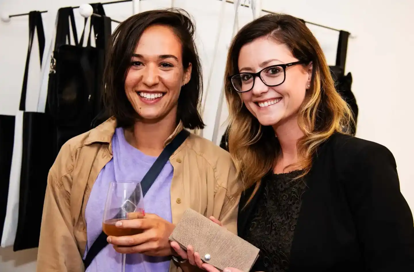 Zwei Frauen beim Networking auf einem Firmenevent Wien, eine hält ein Glas Wein, im Hintergrund schwarze Kleidungsstücke an e