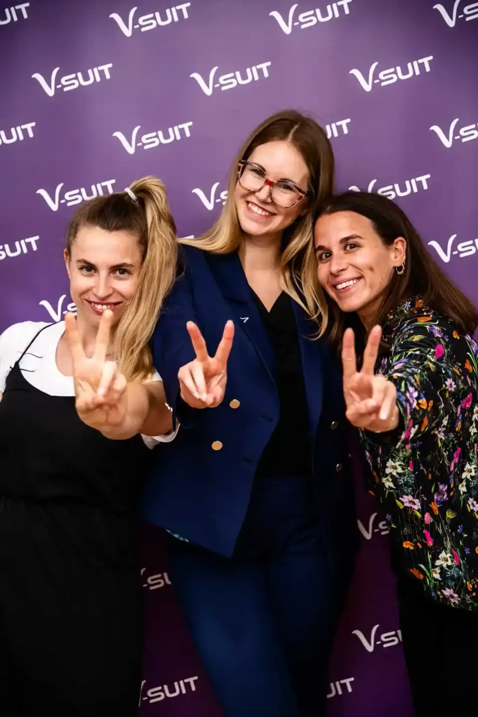 Drei lächelnde Frauen machen Peace-Zeichen vor lila V-SUIT Hintergrund bei einem Firmenevent in Wien