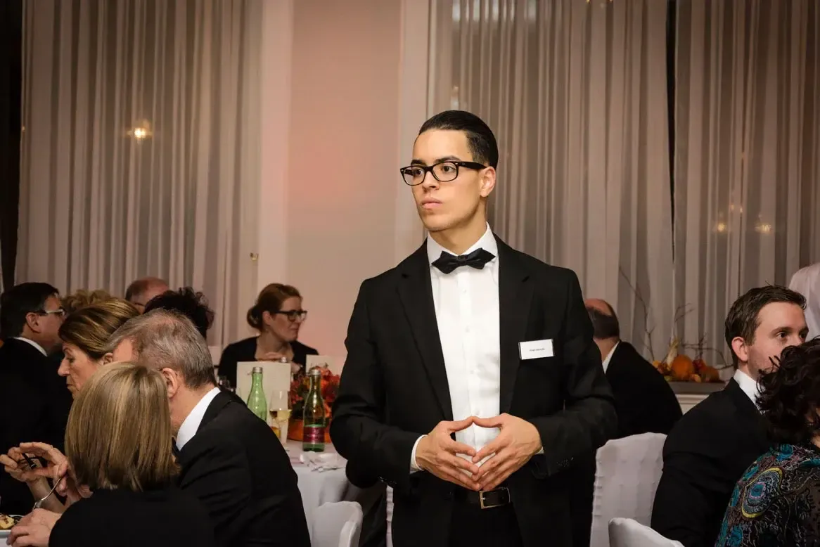 Junger Mann in Smoking und Brille bei Firmenevent Wien, steht zwischen Gästen im eleganten Saal