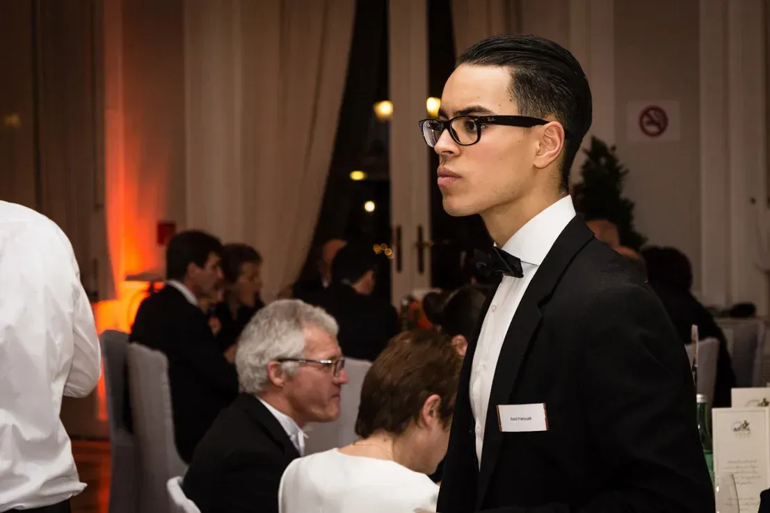 Junger Mann in schwarzem Anzug und Brille bei einem Firmenevent Wien, im Hintergrund Gäste und warme Beleuchtung