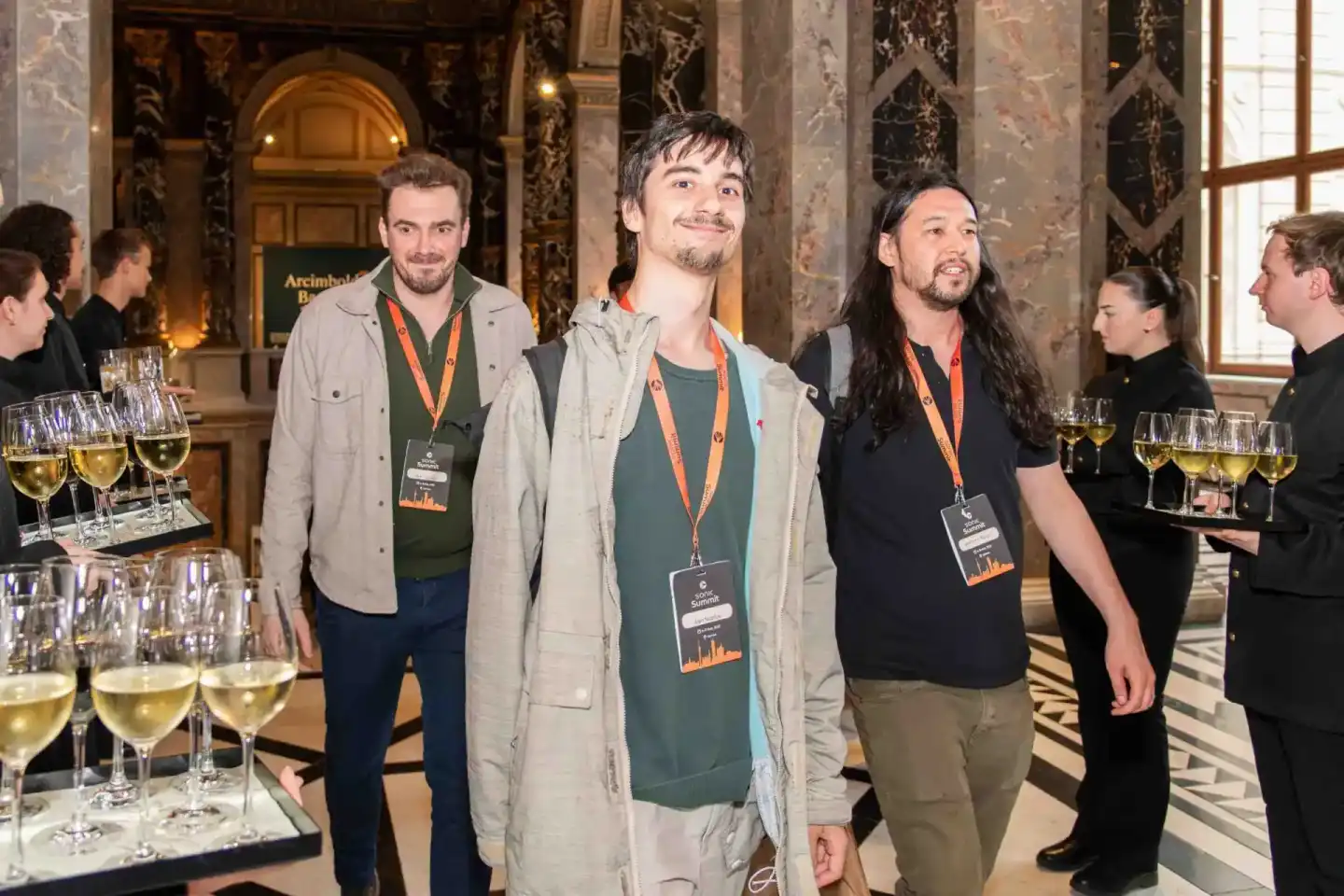 Drei Männer mit Lanyards beim Sonic Summit Firmenevent in Wien, umgeben von Weingläsern in eleganter Halle