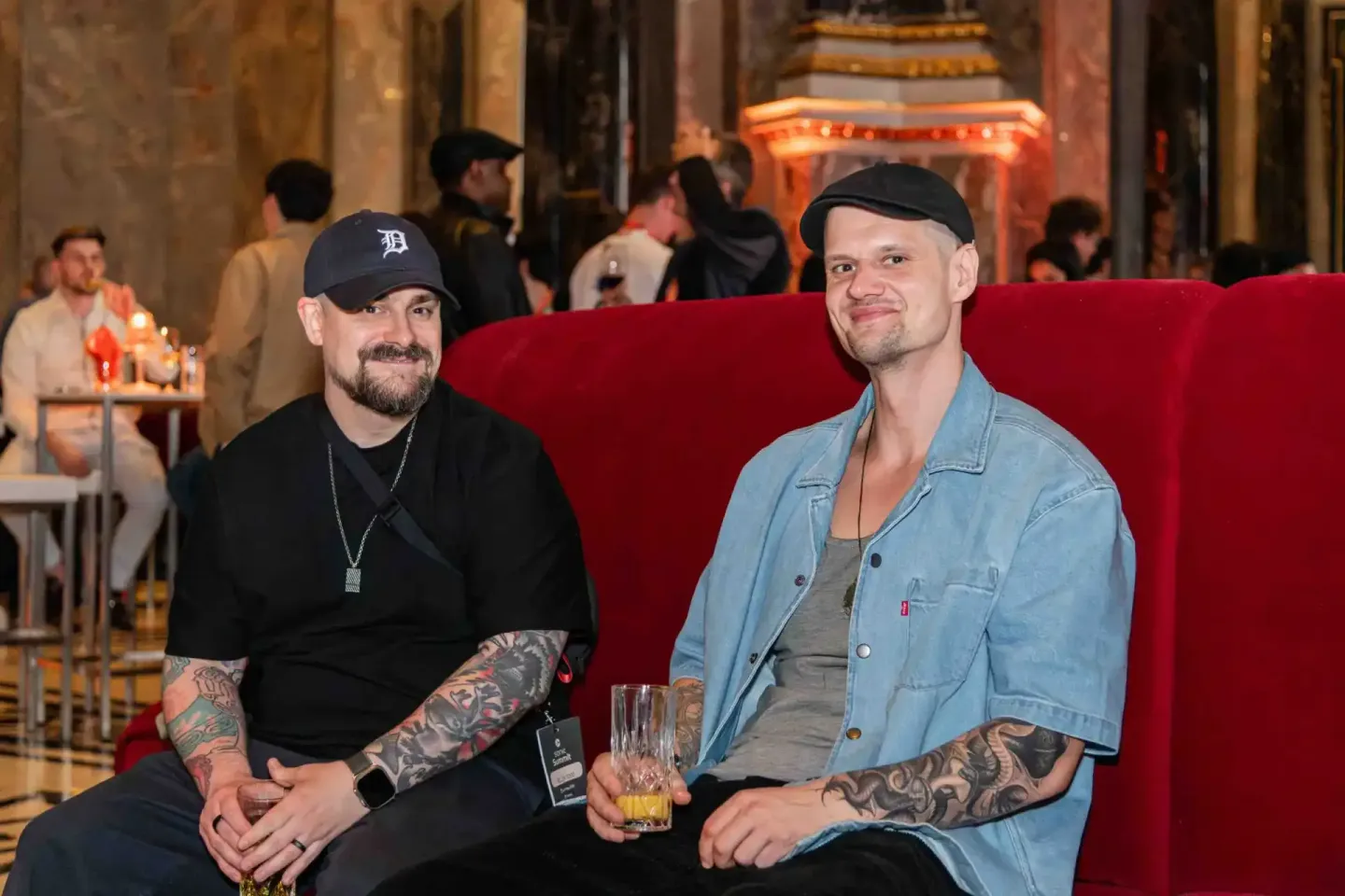 Zwei Männer sitzen auf rotem Sofa bei Sonic Summit Event in Wien, tragen legere Kleidung und halten Getränke, Eventfotografie