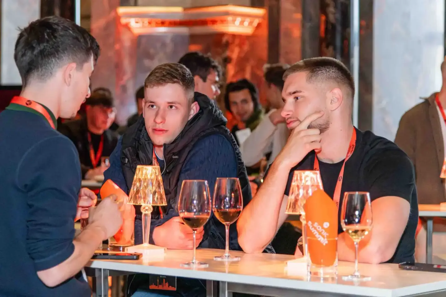 Drei junge Männer in schwarzen Shirts mit orangenen Lanyards unterhalten sich an einem Stehtisch während eines Firmenevent in