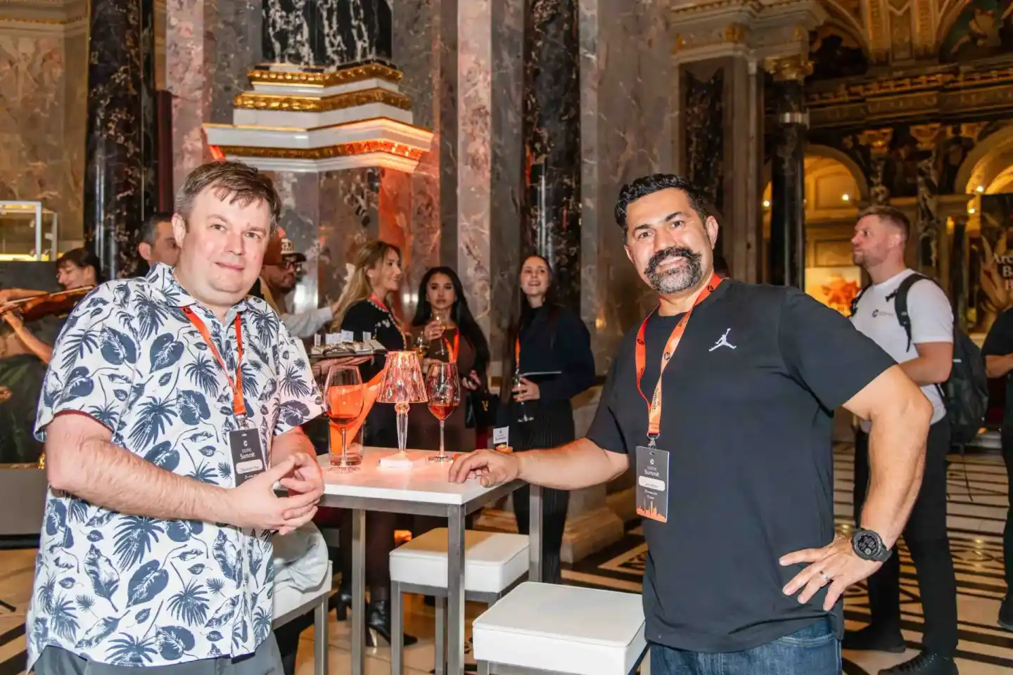 Zwei Männer mit Lanyards stoßen mit Cocktails an, im Hintergrund Gäste in eleganter Halle – Eventfotografie Wien