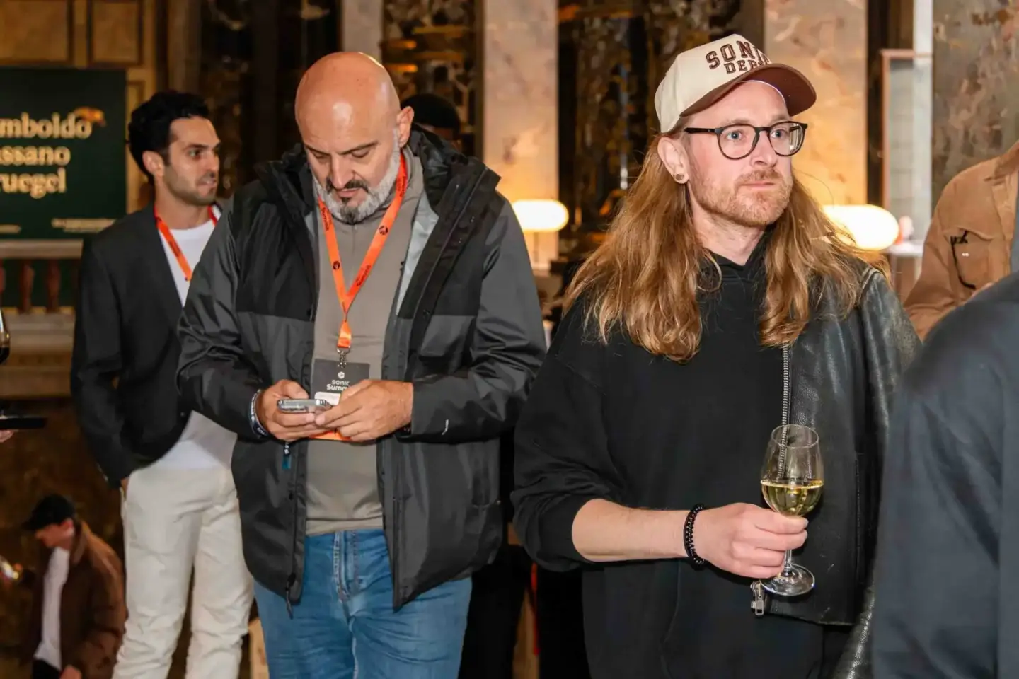 Gäste beim Sonic Summit Event in Wien: Mann mit Langhaar und Brille hält Weinglas, daneben bald Herr mit Lanyard und ID-Badge