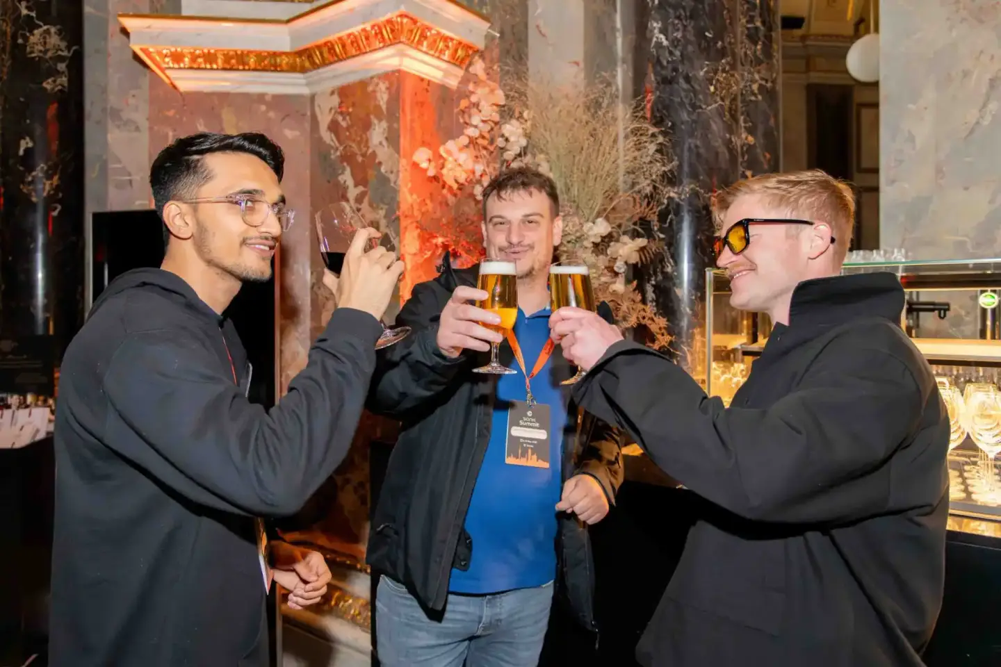 Drei Männer stoßen mit Bierglässern an, im Hintergrund eine orange-weiße Dekoration bei einem Firmenevent in Wien