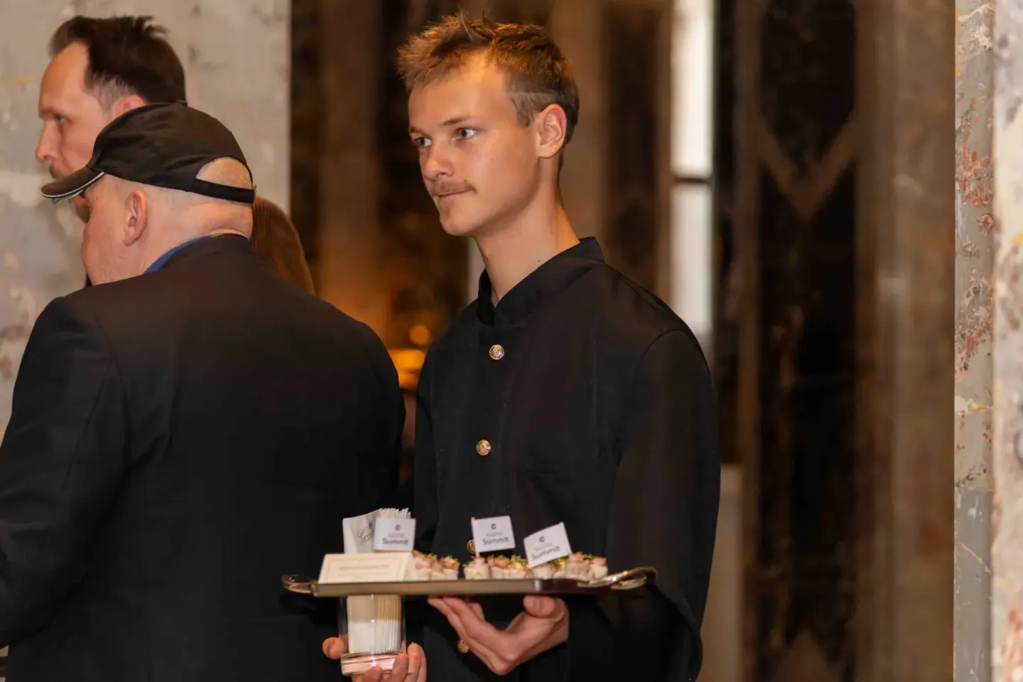 Kellner in schwarzer Uniform serviert Canapés auf Tablett bei Firmenevent Wien, Gast im Hintergrund