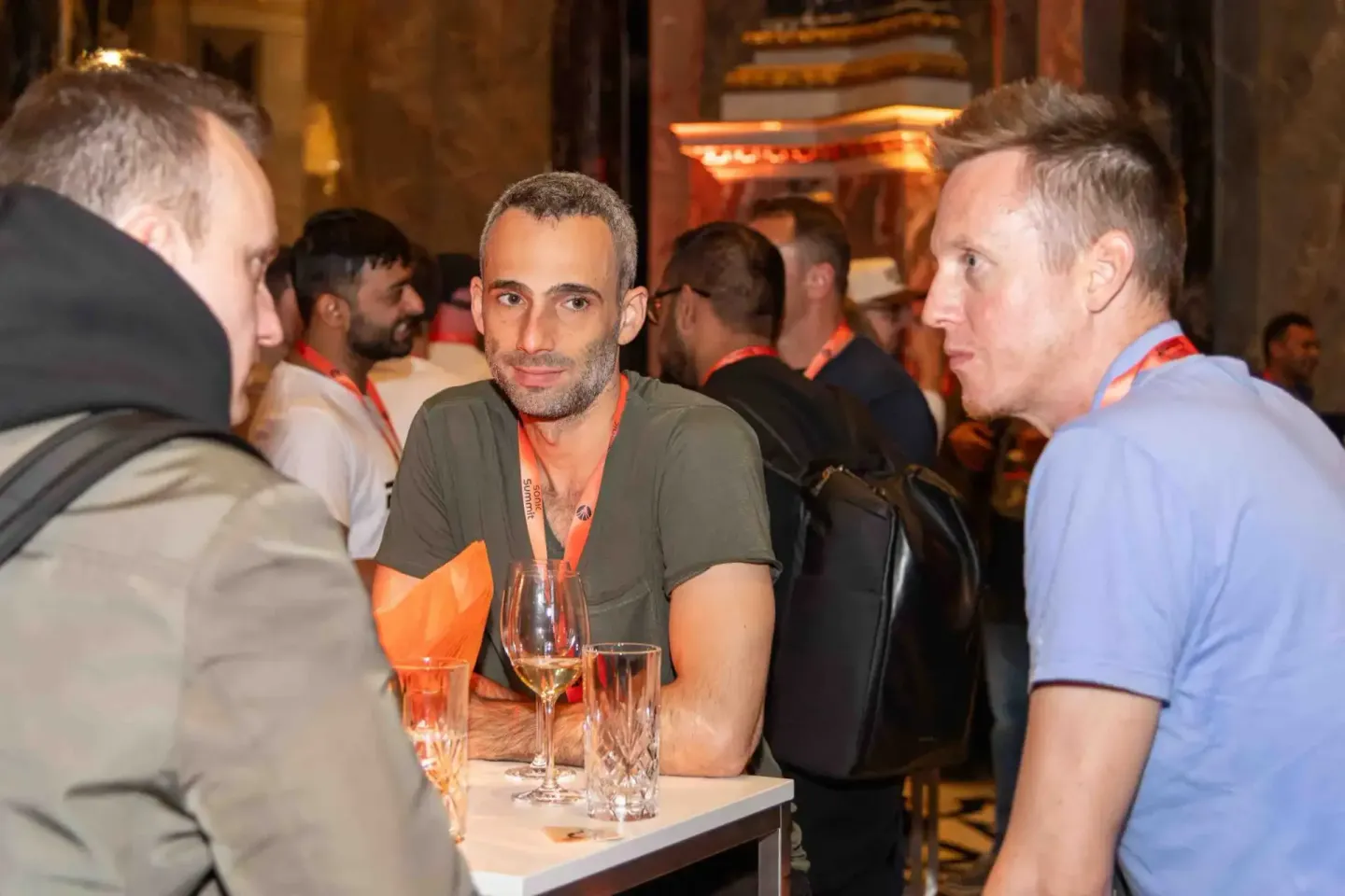 Drei Männer unterhalten sich beim Sonic Summit Firmenevent in Wien, einer trägt ein oranges Lanyband, Getränke auf dem Tisch