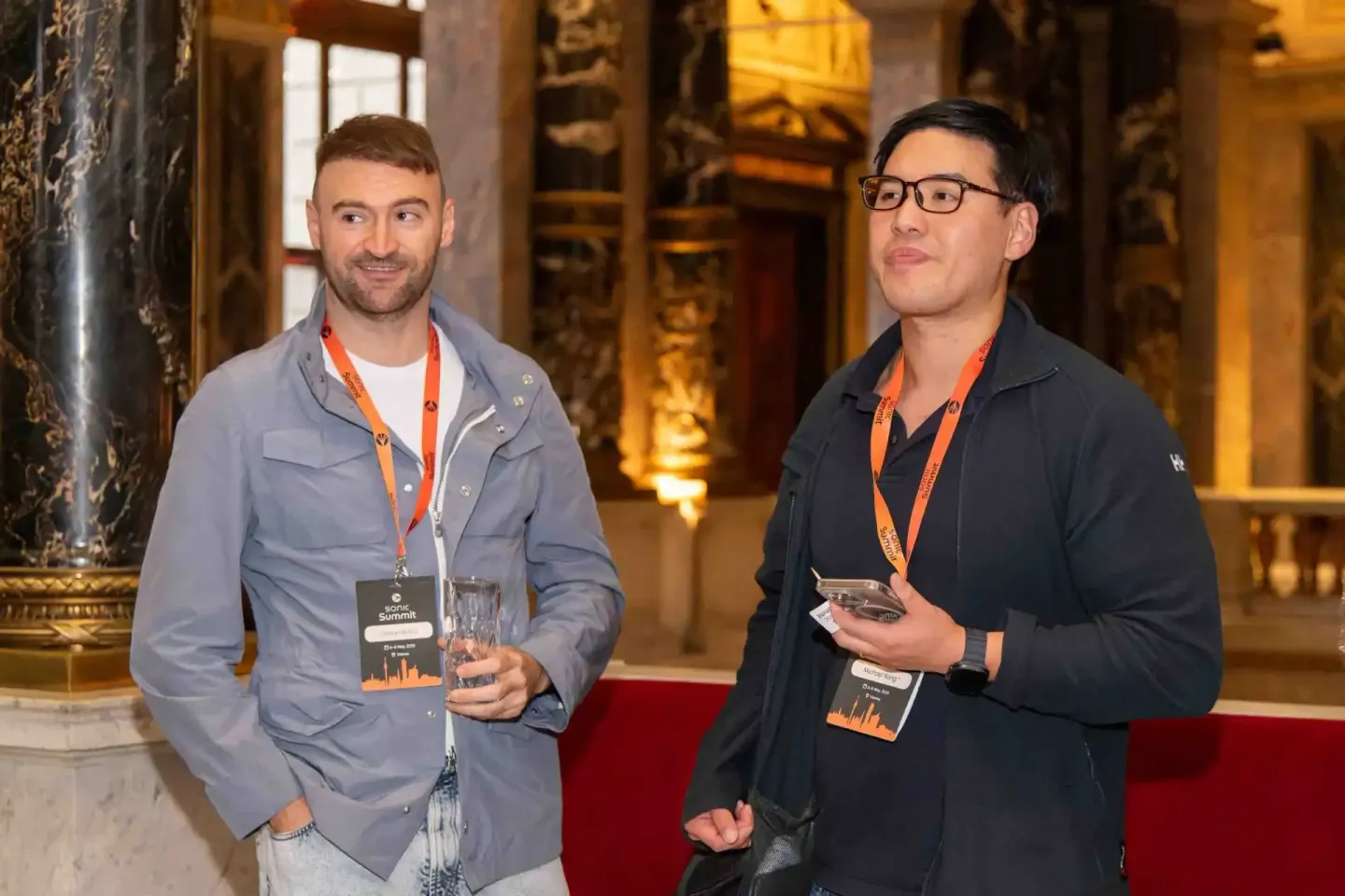 Zwei Teilnehmer beim Sonic Summit mit Lanyards und Getränken in eleganter Veranstaltungshalle, professionelle Eventfotografie