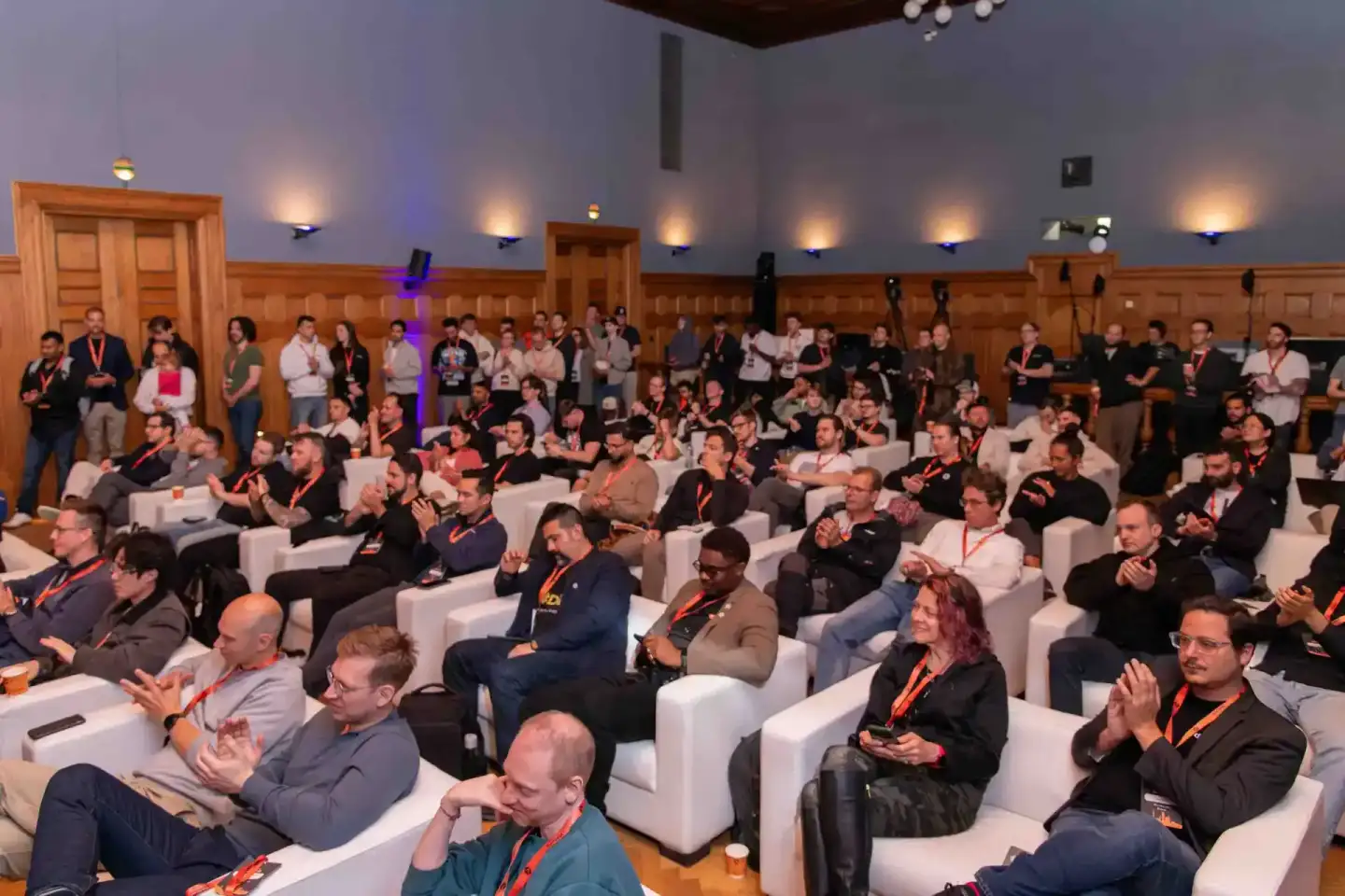 Große Konferenzveranstaltung im eleganten Holzsaal mit etwa 100 Gästen in weißen Sesseln, viele tragen rote Lanyards. Profess