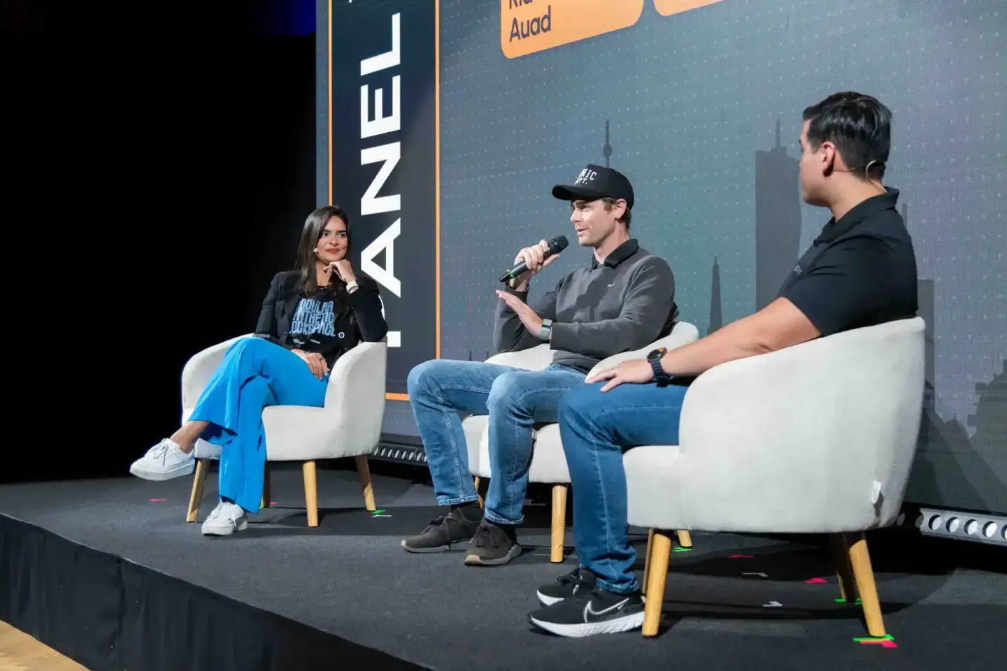 Drei Personen sitzen auf weißen Sesseln auf einer Bühne beim Sonic Summit Event. Der Mann in der Mitte hält ein Mikrofon. Eve