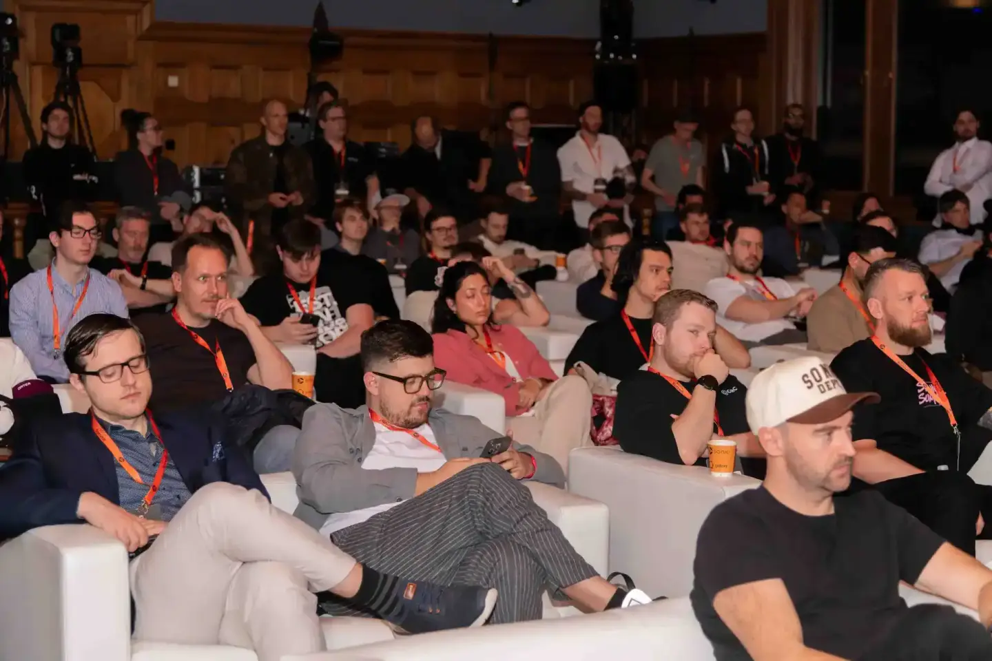Publikum sitzt auf weißen Sesseln während einer Konferenzveranstaltung, viele Teilnehmer tragen orangefarbene Lanyards beim S