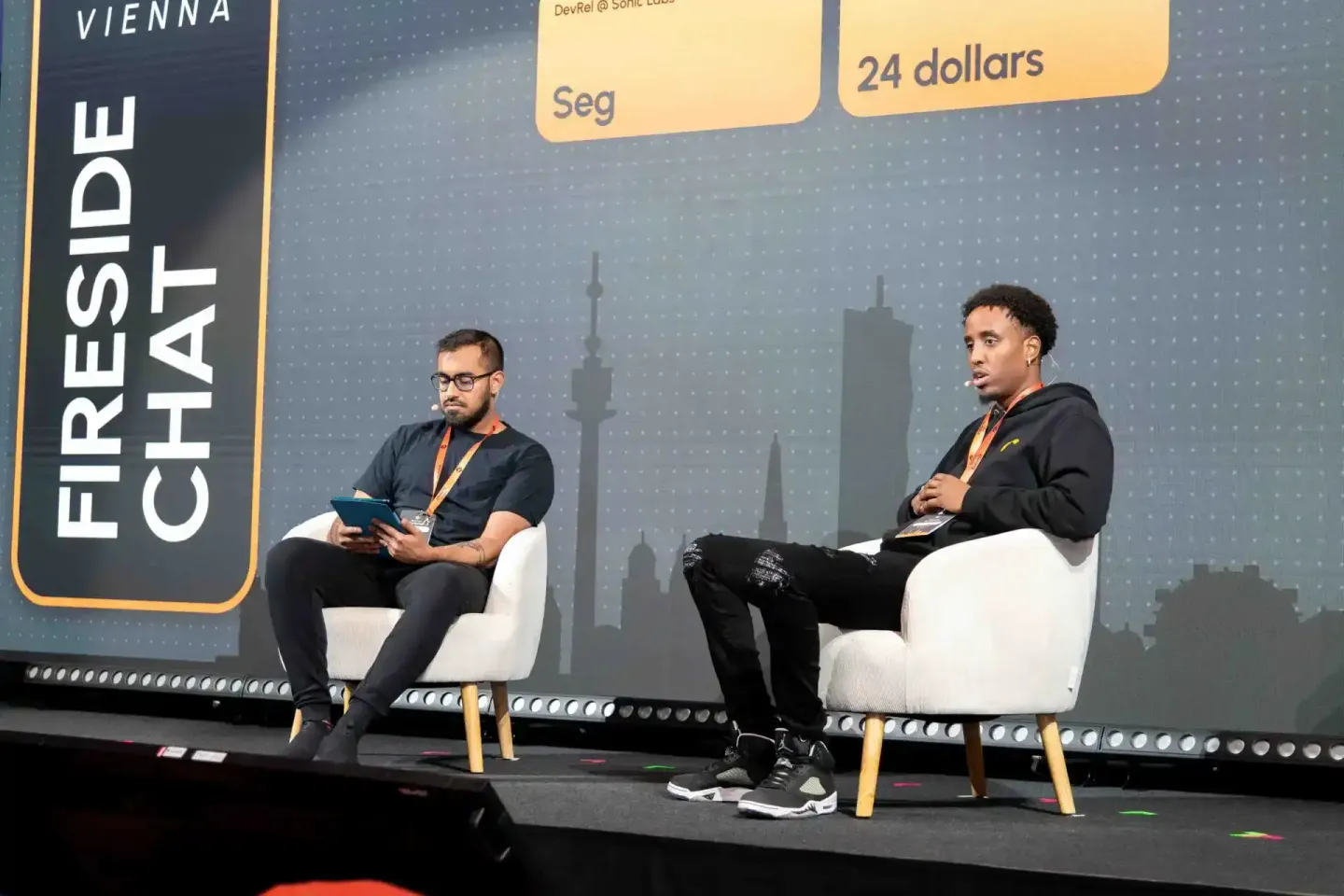 Zwei Sprecher beim Sonic Summit in Wien sitzen auf weißen Sesseln auf der Bühne und diskutieren vor einer Skyline-Projektion