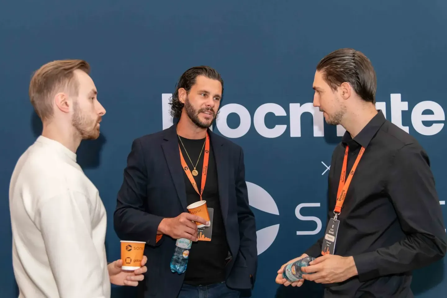 Drei Männer im Gespräch beim Sonic Summit Event mit Getränken und Lanyards vor blauer Wand