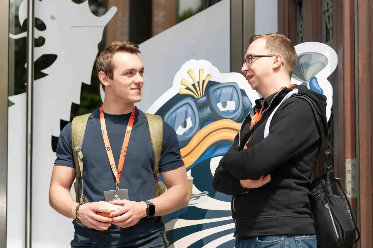 Zwei Teilnehmer im Gespräch beim Sonic Summit Event in Wien, einer trägt ein Lanyardabzeichen, im Hintergrund bunte Event-Gra