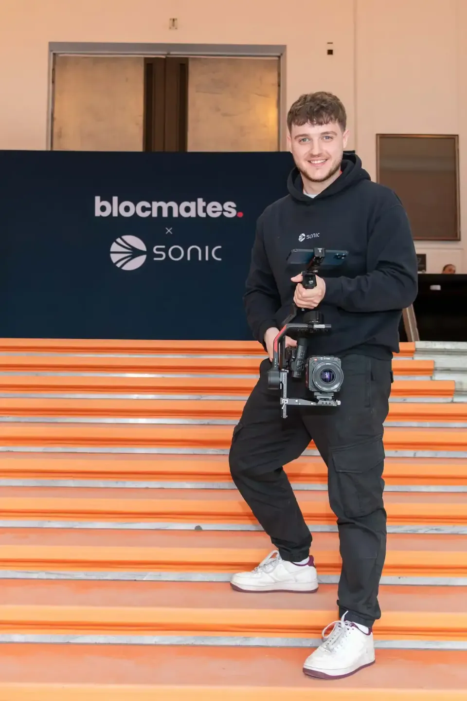 Junger Mann mit Kamera-Gimbal vor Blocmates und Sonic Logo auf orangen Stufen beim Sonic Summit Event