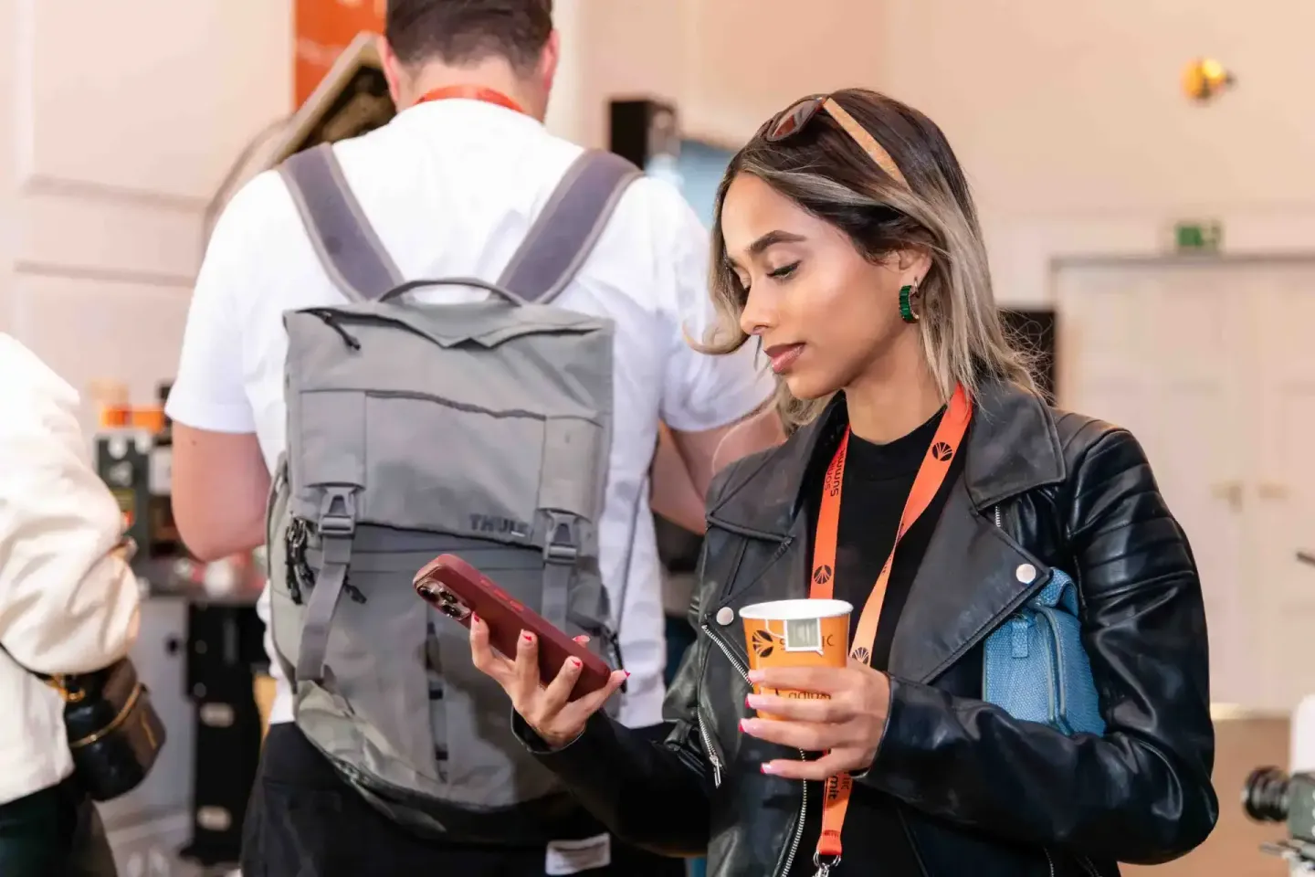 Besucherin mit Getränk und Smartphone beim Sonic Summit Event, im Hintergrund Mann mit grauem Thule-Rucksack