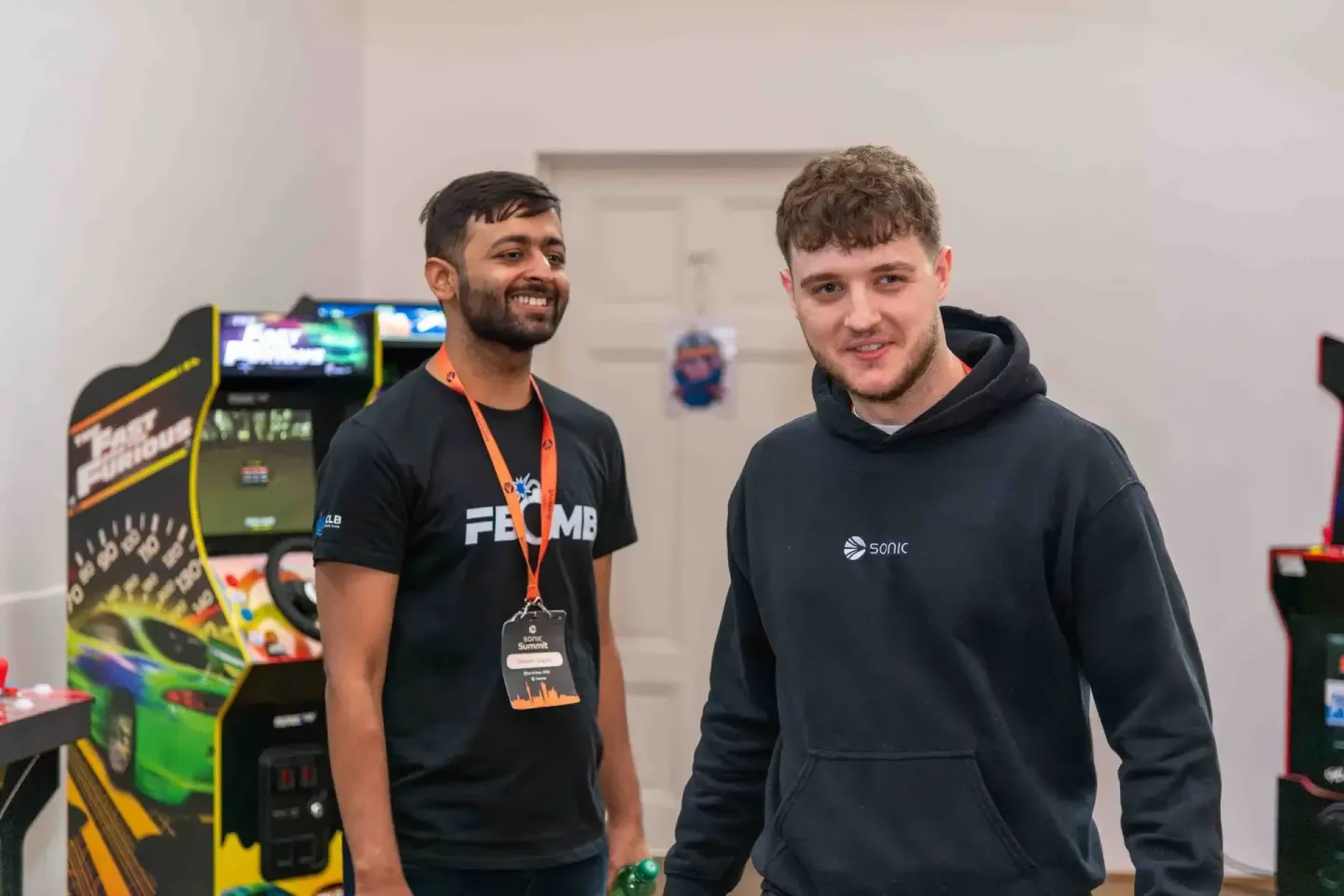 Zwei Männer beim Sonic Summit Event in Wien, einer trägt ein FRAME-Shirt mit Badge, im Hintergrund Arcade-Automaten