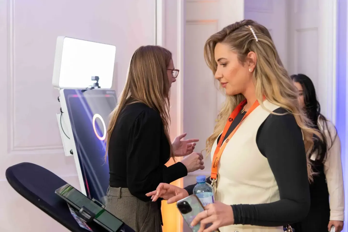 Zwei Frauen im Gespräch bei einem Firmenevent Wien, eine trägt eine orange Lanyard, im Hintergrund eine digitale Displaystati