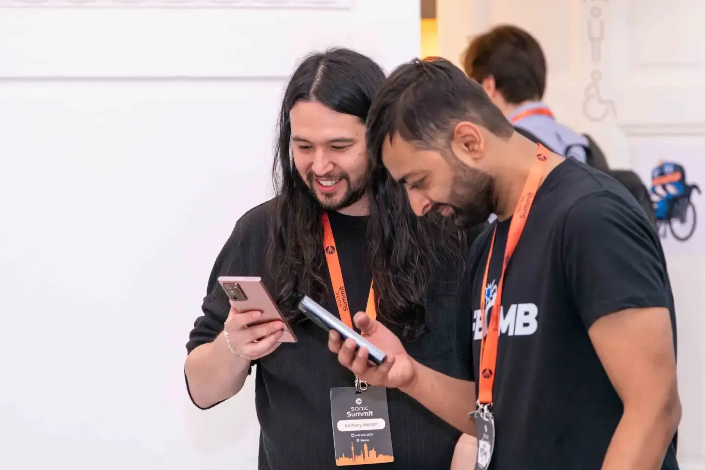 Drei Besucher mit Badges beim Sonic Summit Event schauen gemeinsam auf ein Smartphone, Eventfotograf dokumentiert die Atmosph