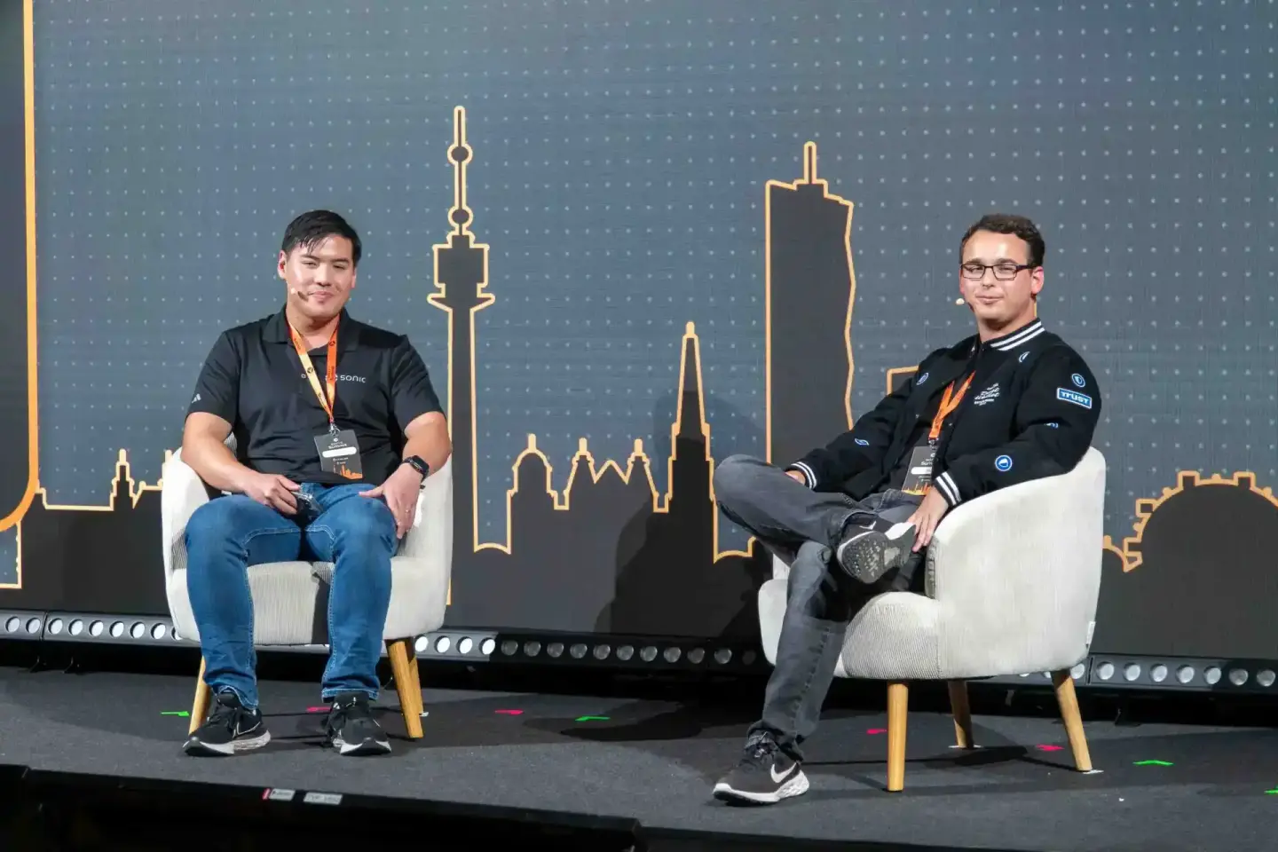 Zwei Sprecher sitzen sich auf der Bühne des Sonic Summit gegenüber. Im Hintergrund ist eine Skyline aus goldenen Gebäudeumris