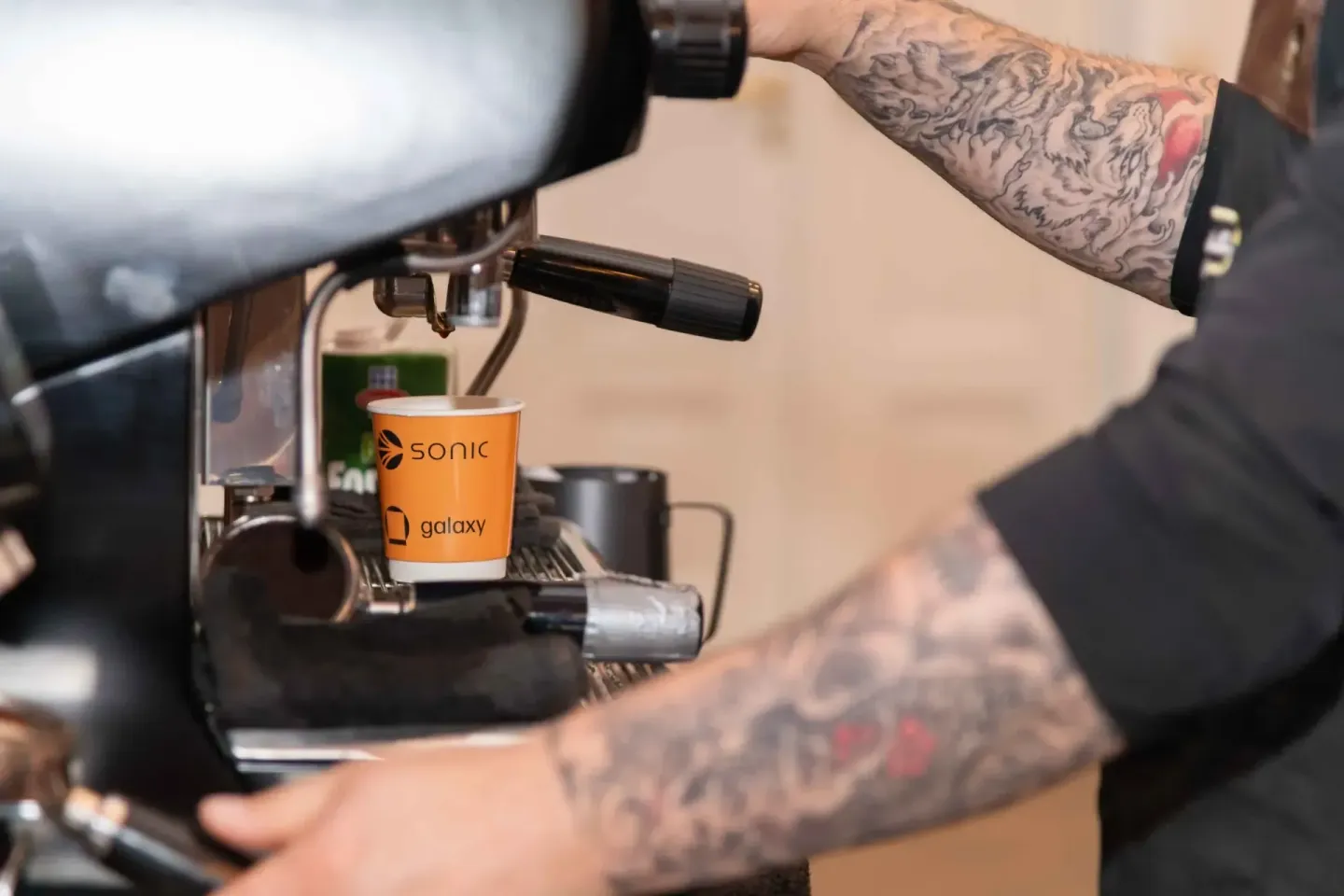 Barista mit Tätowierungen bedient professionell eine Espresso-Maschine und füllt einen orange bedruckten Sonic-Galaxy-Becher 