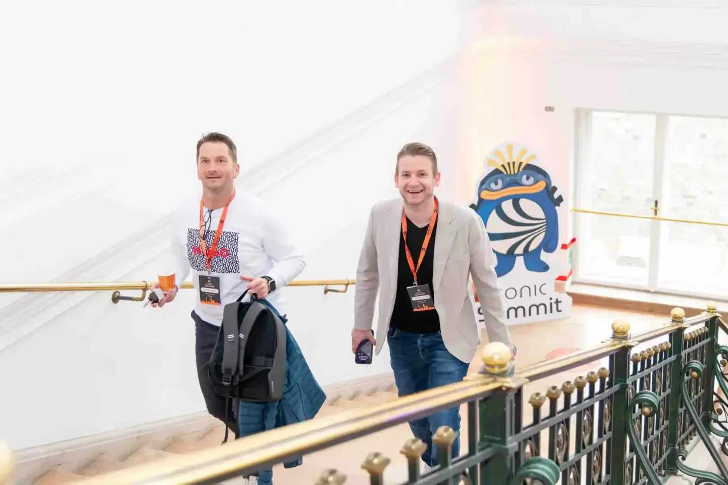 Zwei Männer mit Konferenzausweisen laufen lächelnd durch ein modernes Treppenhaus beim Sonic Summit in Wien – professionelle 
