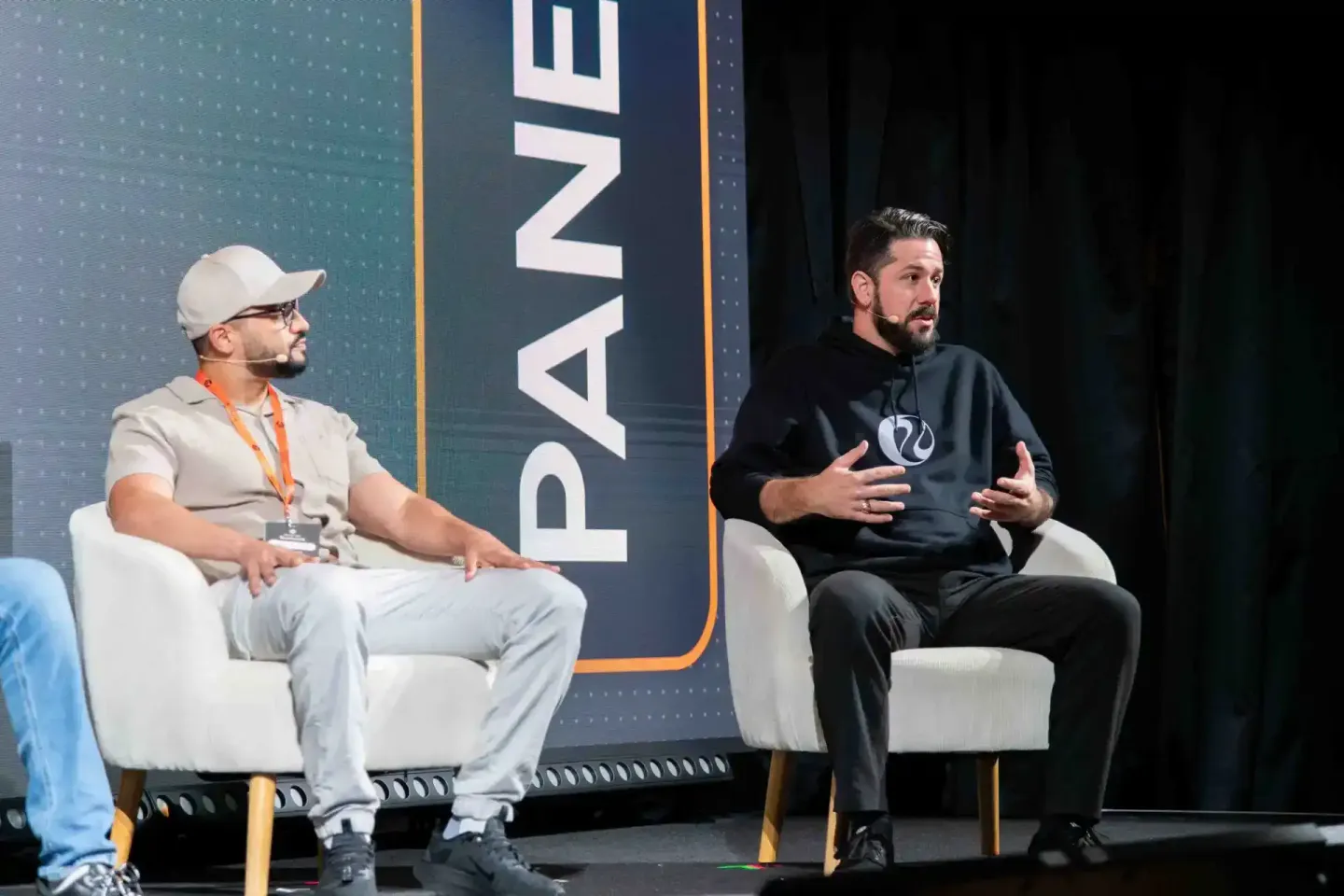 Zwei Sprecher sitzen auf der Bühne beim Sonic Summit Event in Wien. Ein Mann in beigem Hemd und Cap moderiert das Gespräch mi