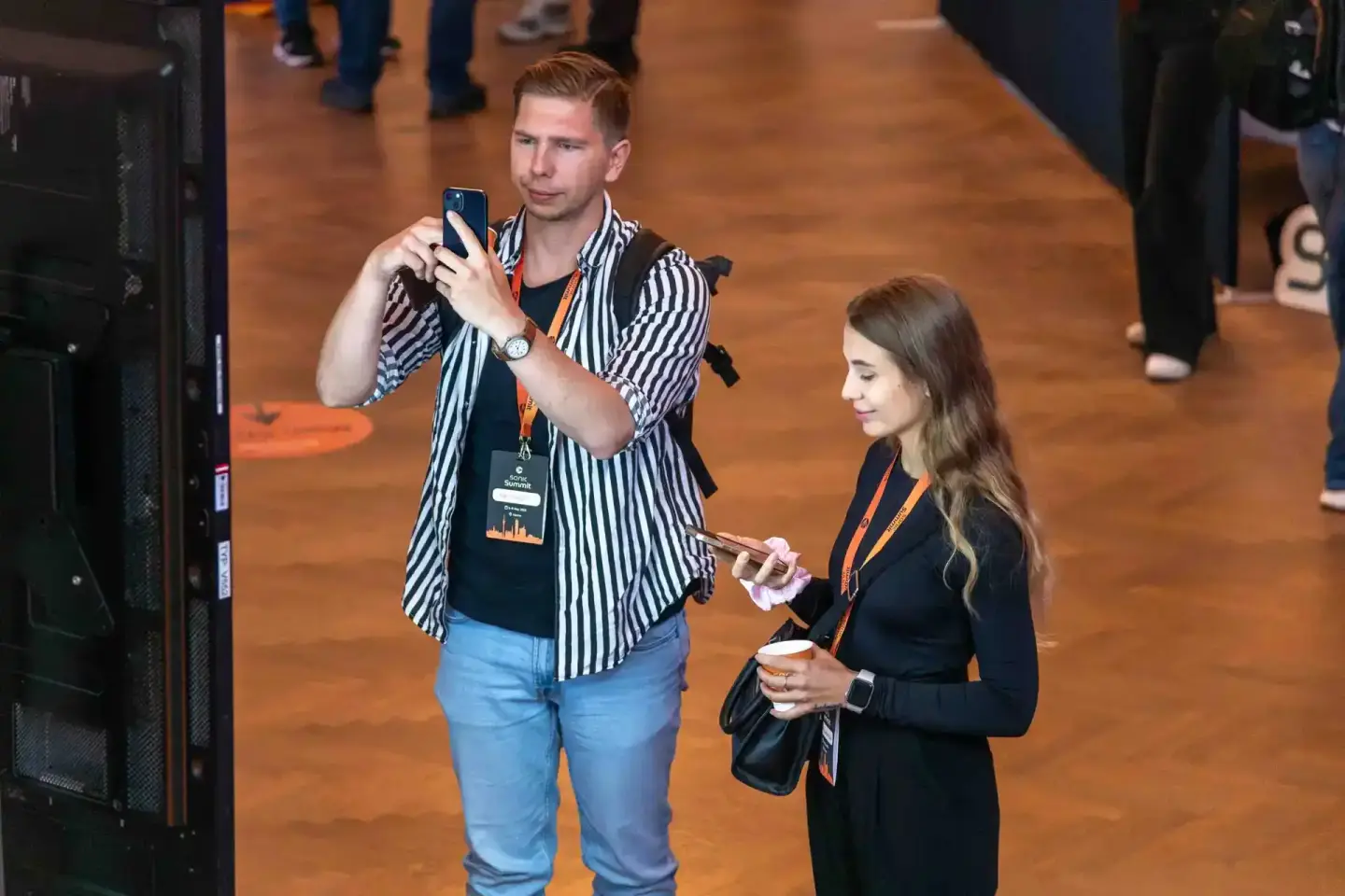 Eventfotograf dokumentiert Besucher beim Sonic Summit: Mann mit Smartphone und Frau mit Lanyards im Gespräch auf der Veransta