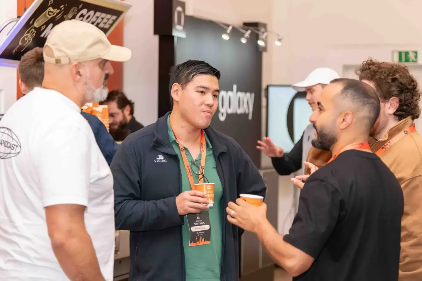 Teilnehmer beim Sonic Summit Event in Wien im Gespräch mit einem Eventfotograf, der ein Getränk hält und ein Namensschild trä