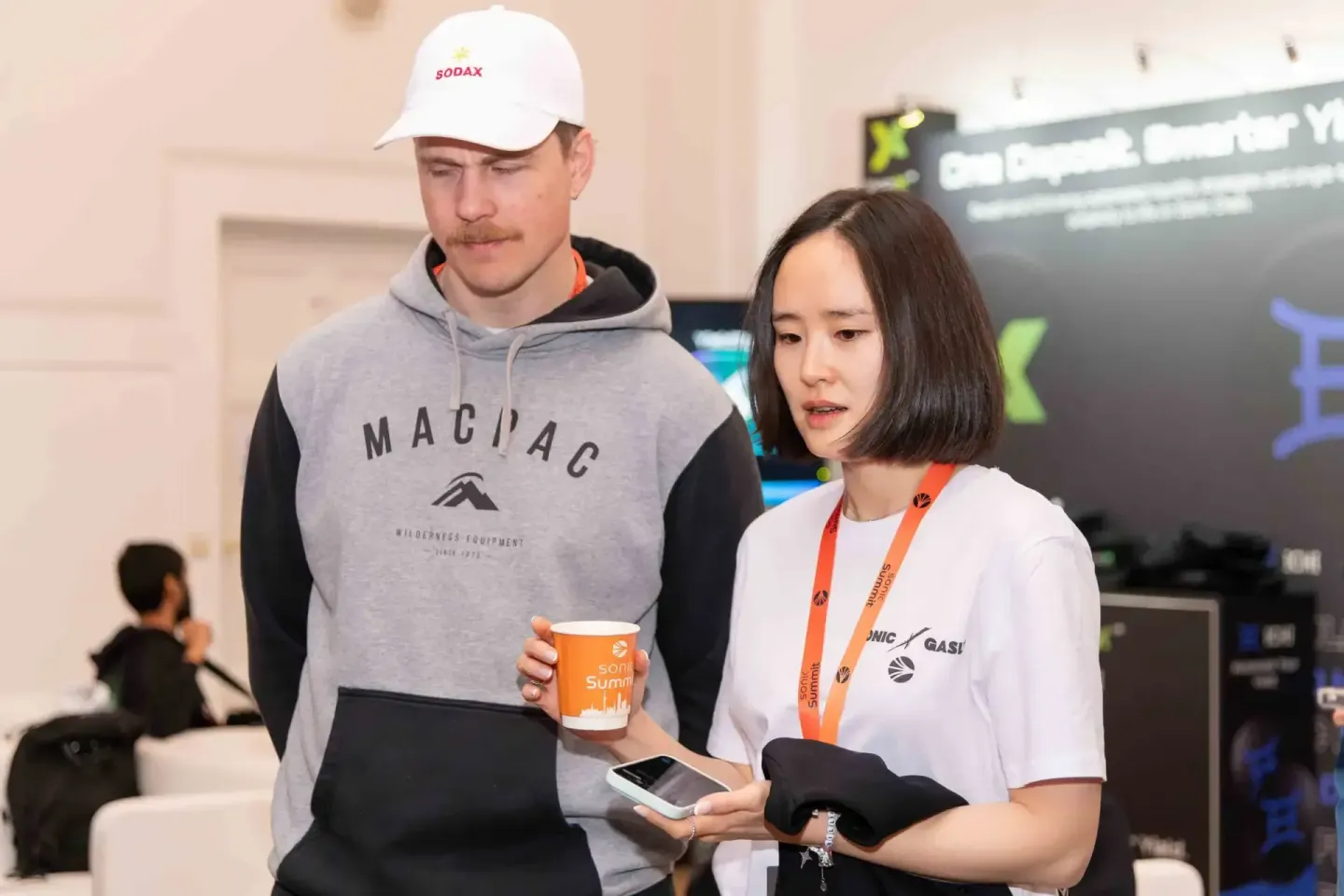 Eventfotograf dokumentiert Gespräch zwischen zwei Personen beim Sonic Summit – Mann mit SODAX-Cap und Frau mit orange Lanyard