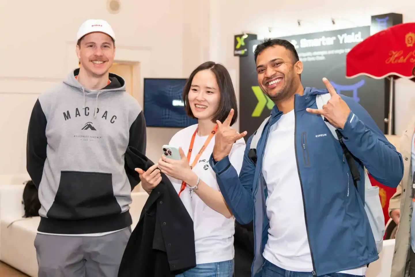 Drei lachende Personen bei einem Firmenevent in Wien, die Mann in Mütze und Hoodie, eine Frau mit Lanyards hält Smartphone, M