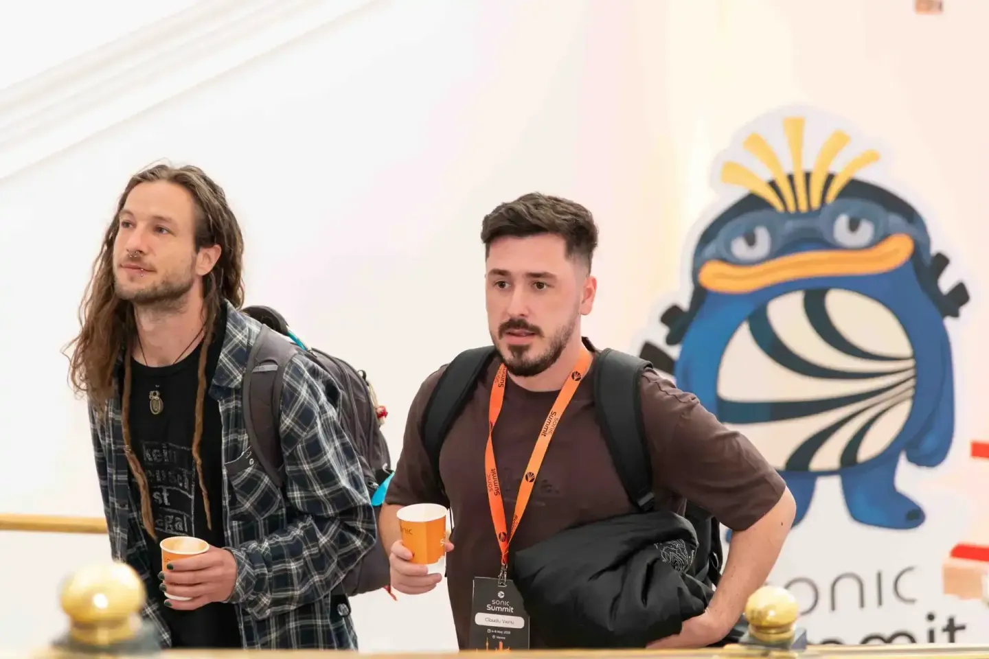 Zwei Männer beim Sonic Summit Event in Wien, einer mit langen Haaren und kariertem Hemd, einer mit Namensschild, vor blauem M