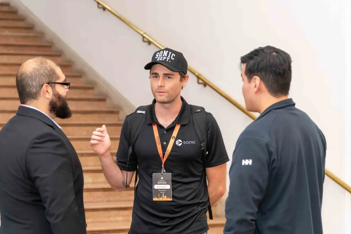 Drei Männer im Gespräch bei der Sonic Summit. Der mittlere Mann trägt ein schwarzes Sonic-Polo-Shirt und Accreditation Badge.