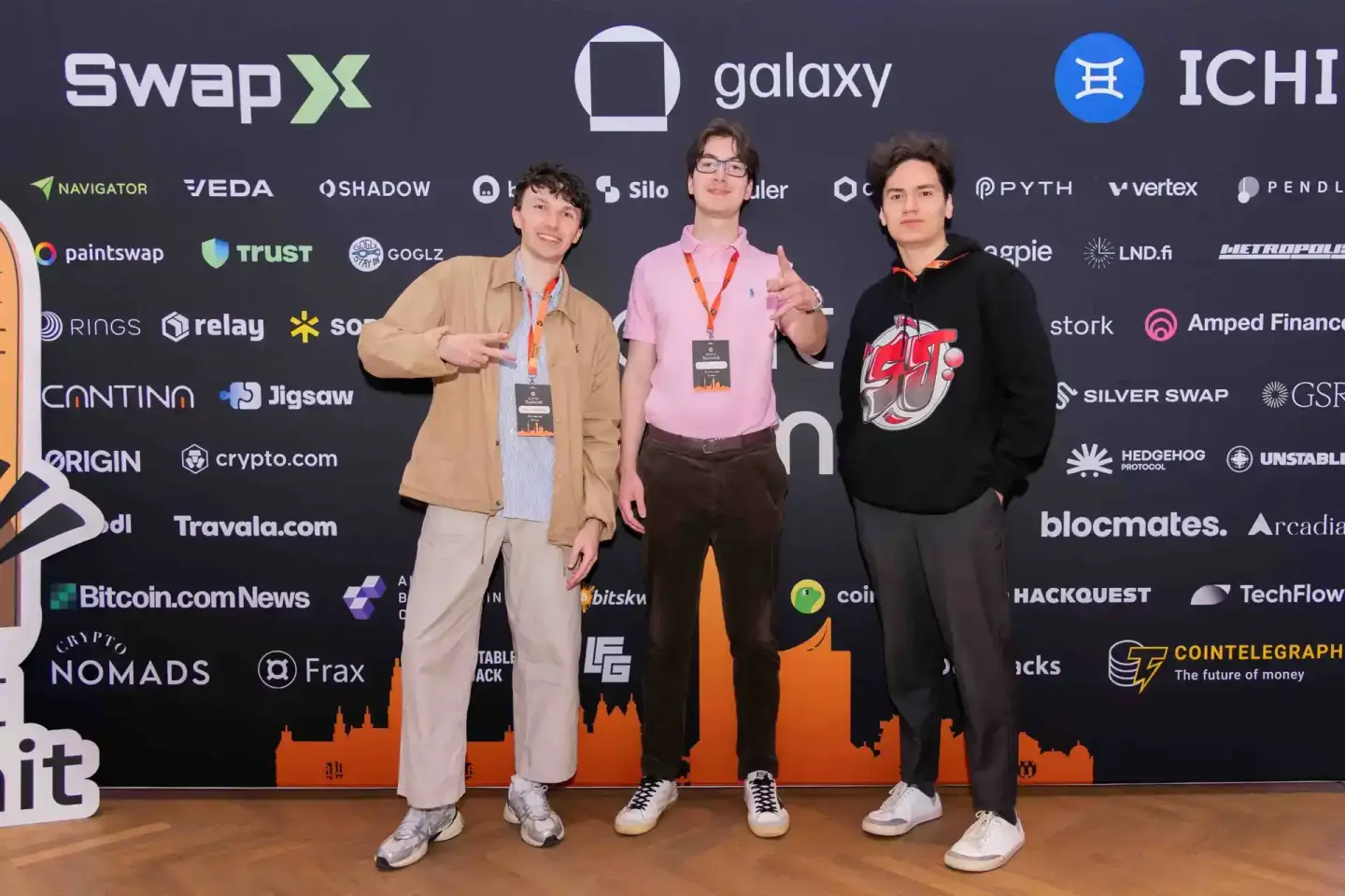 Drei Personen mit Event-Badges posieren vor einer großen Bühne mit Sponsor-Logos bei einem Tech-Summit – professionelle Event