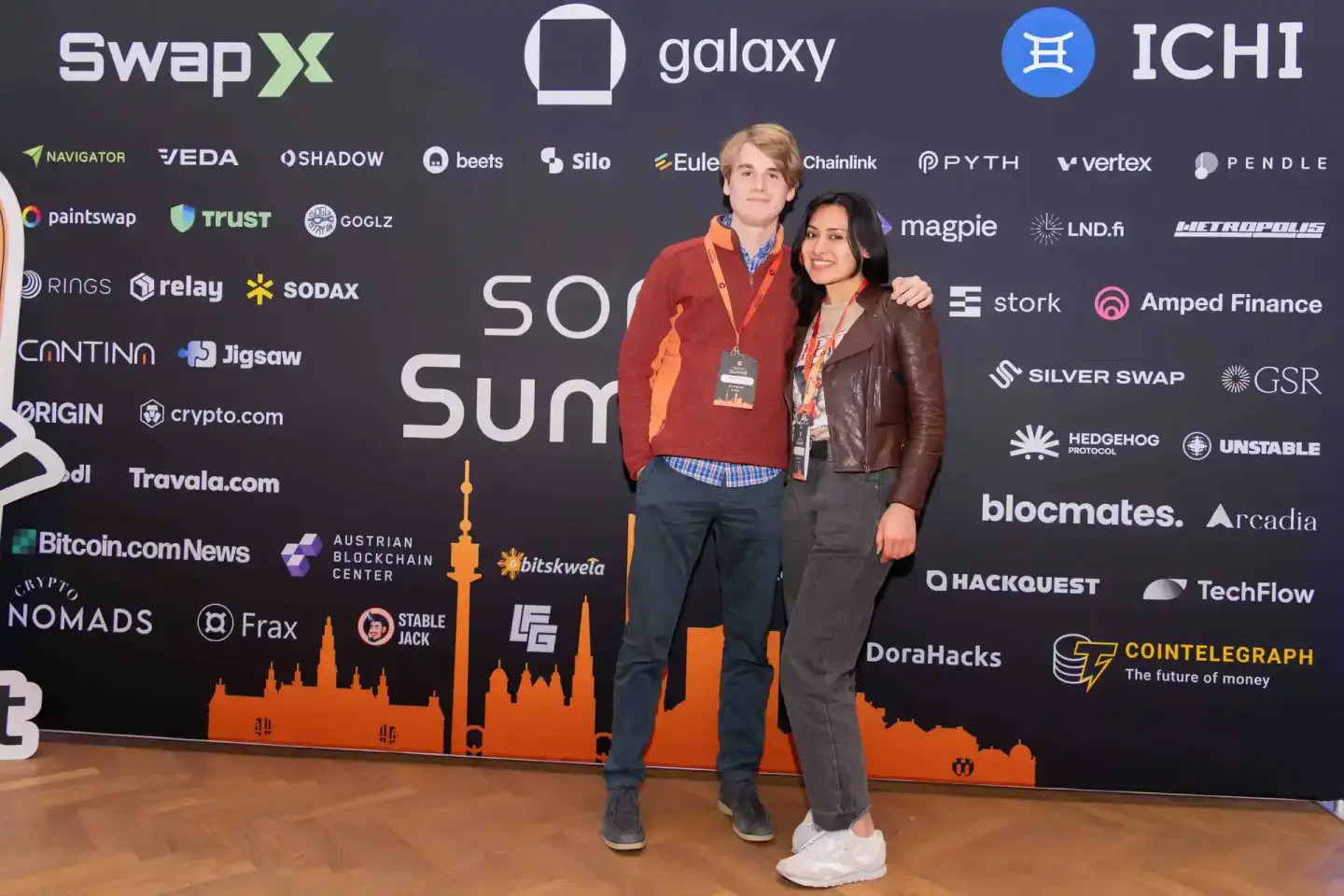Zwei Personen posieren vor einer dunklen Bühnenkulisse mit Eventlogos. Professionelle Eventfotografie Wien.