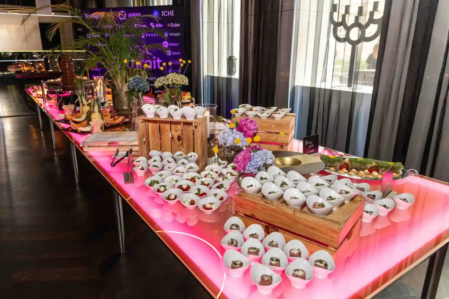 Elegante Event-Catering-Station mit neon-pink beleuchtetem Tisch, weißen Porzellanschüsseln mit Speisen, Blumendekoration und