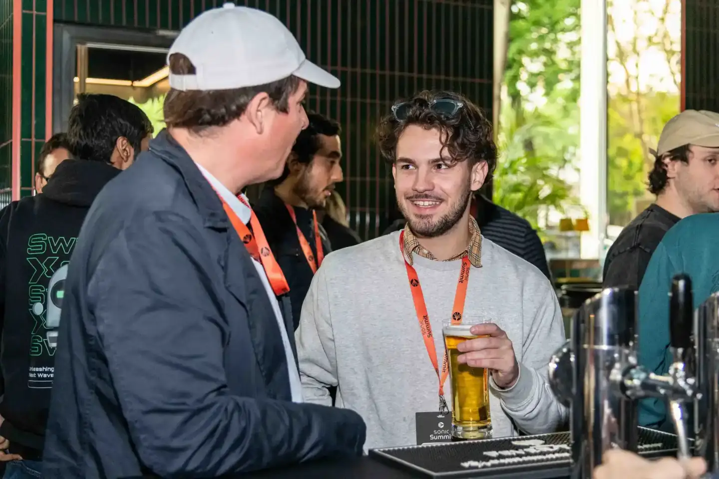 Zwei Männer im Gespräch bei der Sonic Summit Veranstaltung. Der Mann rechts trägt ein rotes Lanyard und hält ein Bier. Eventf