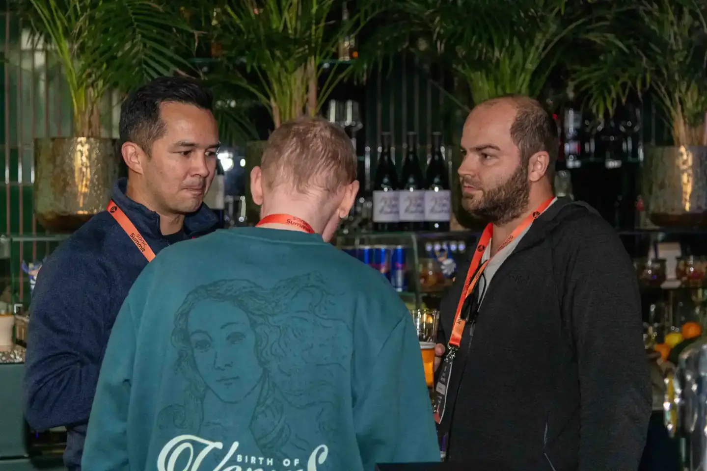 Drei Männer im Gespräch bei einem Firmenevent Wien, zwei tragen orange Lanyards, im Hintergrund grüne Pflanzen und eine Bar