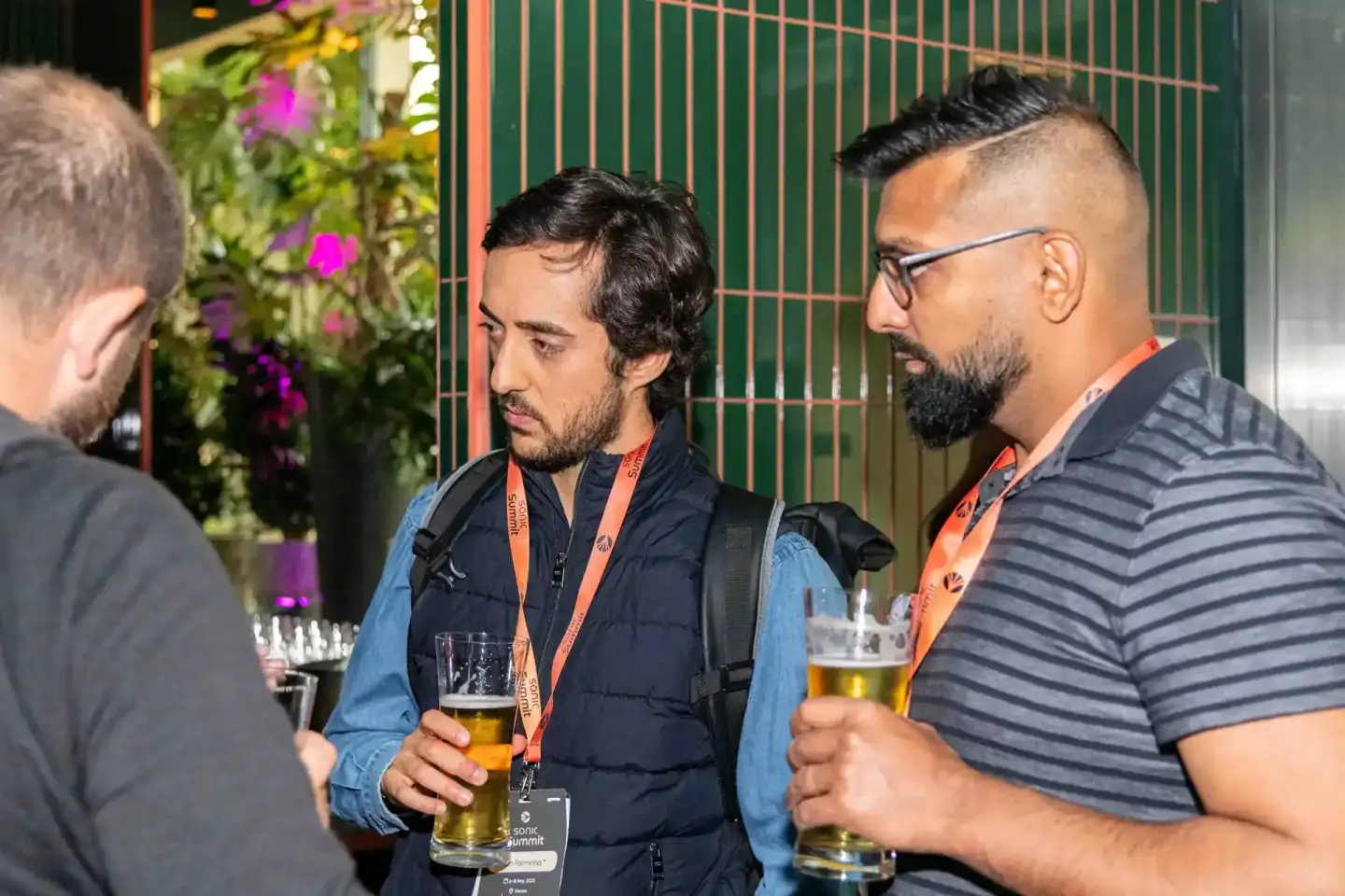 Drei Männer im Gespräch beim Sonic Summit Event in Wien, zwei tragen orange Lanyards und halten Bierglӓser