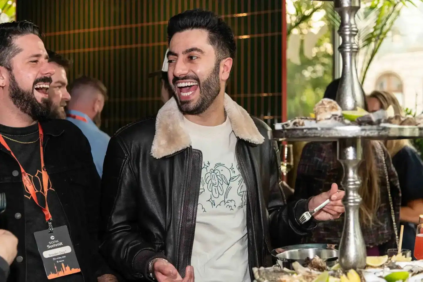 Lachender Mann mit Bart in schwarzer Lederjacke beim Sonic Summit Event in Wien, umgeben von Gästen und Dekorationen