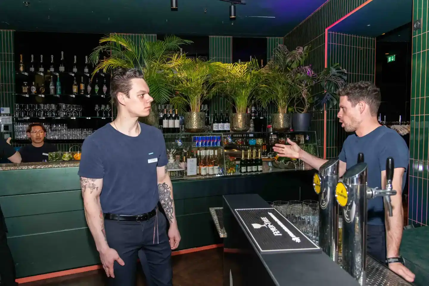 Zwei Männer unterhalten sich an der Bar eines modernen Clubs mit grünen Neonleuchten und Pflanzen - Eventfotografie Wien
