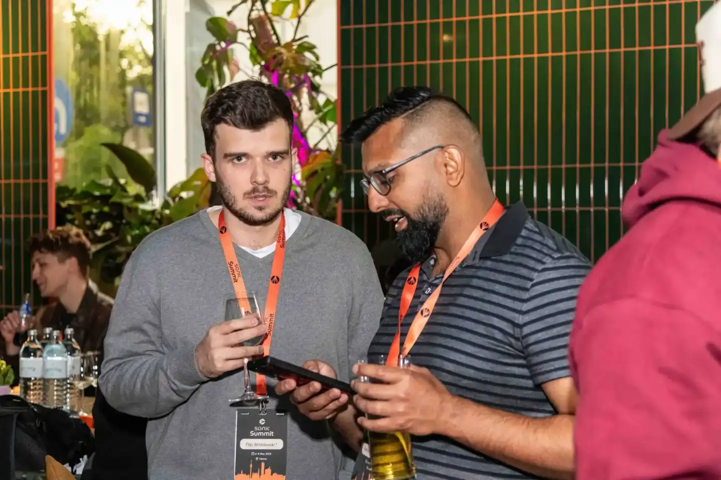 Zwei Besucher mit Lanyards beim Sonic Summit Event in Wien, einer hält ein Smartphone, im Hintergrund grüne Wand und Pflanzen