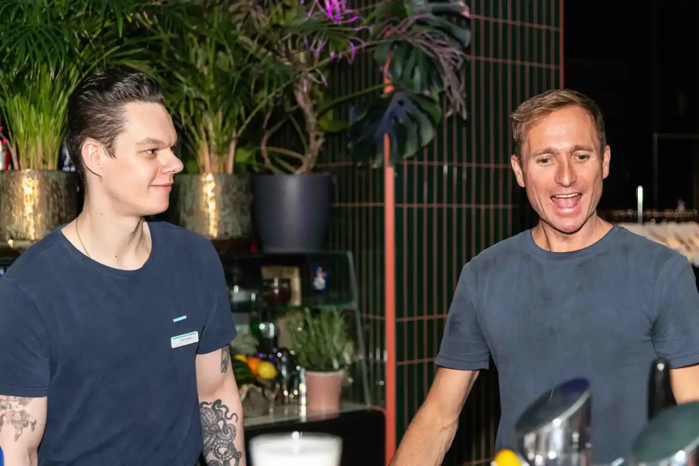 Zwei Männer in dunklen T-Shirts unterhalten sich auf einem Firmevent in Wien, im Hintergrund grüne Pflanzen und moderne Dekor