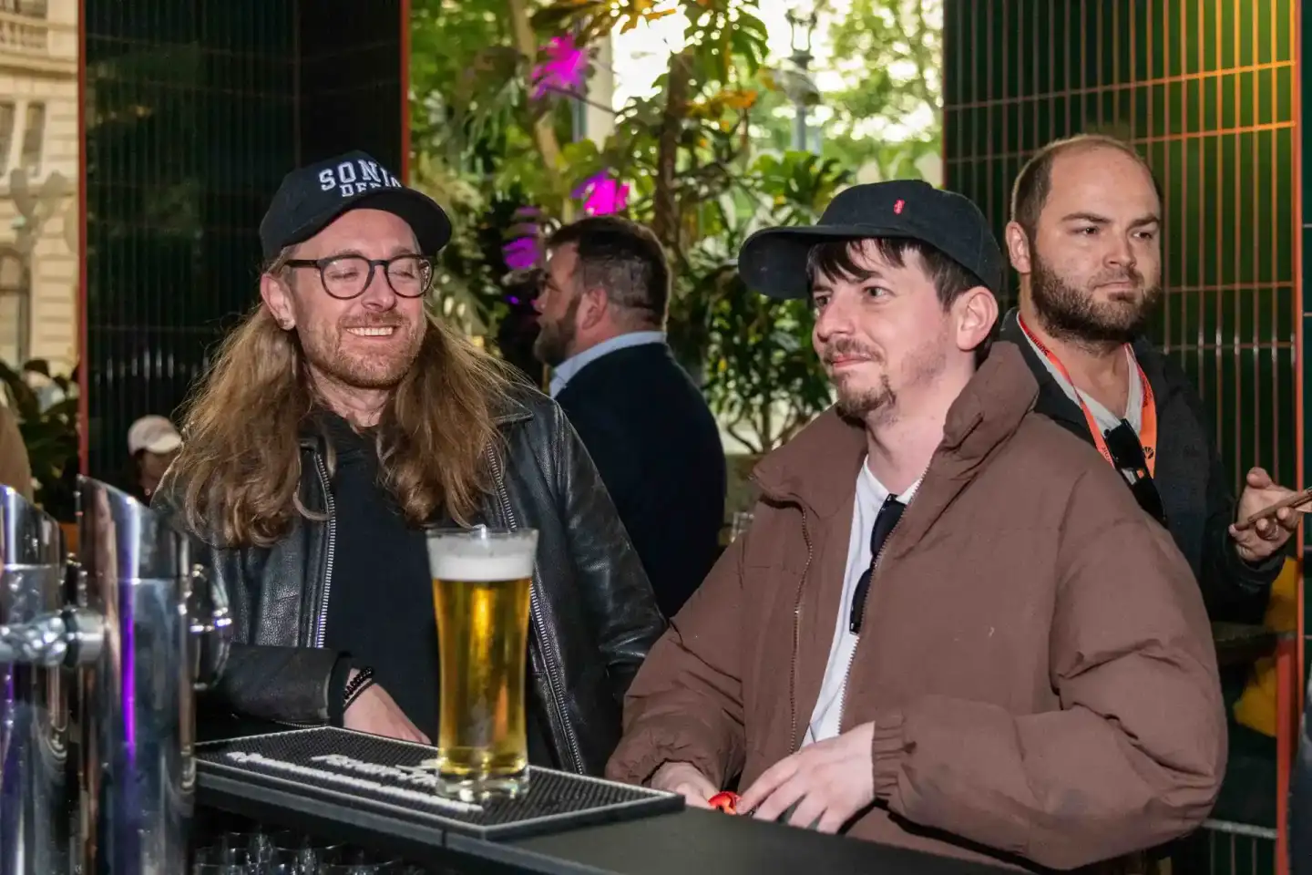 Drei Männer an einer Bar beim Sonic Summit Event in Wien, einer mit Bier in der Hand, im Hintergrund grüne Pflanzen und ander