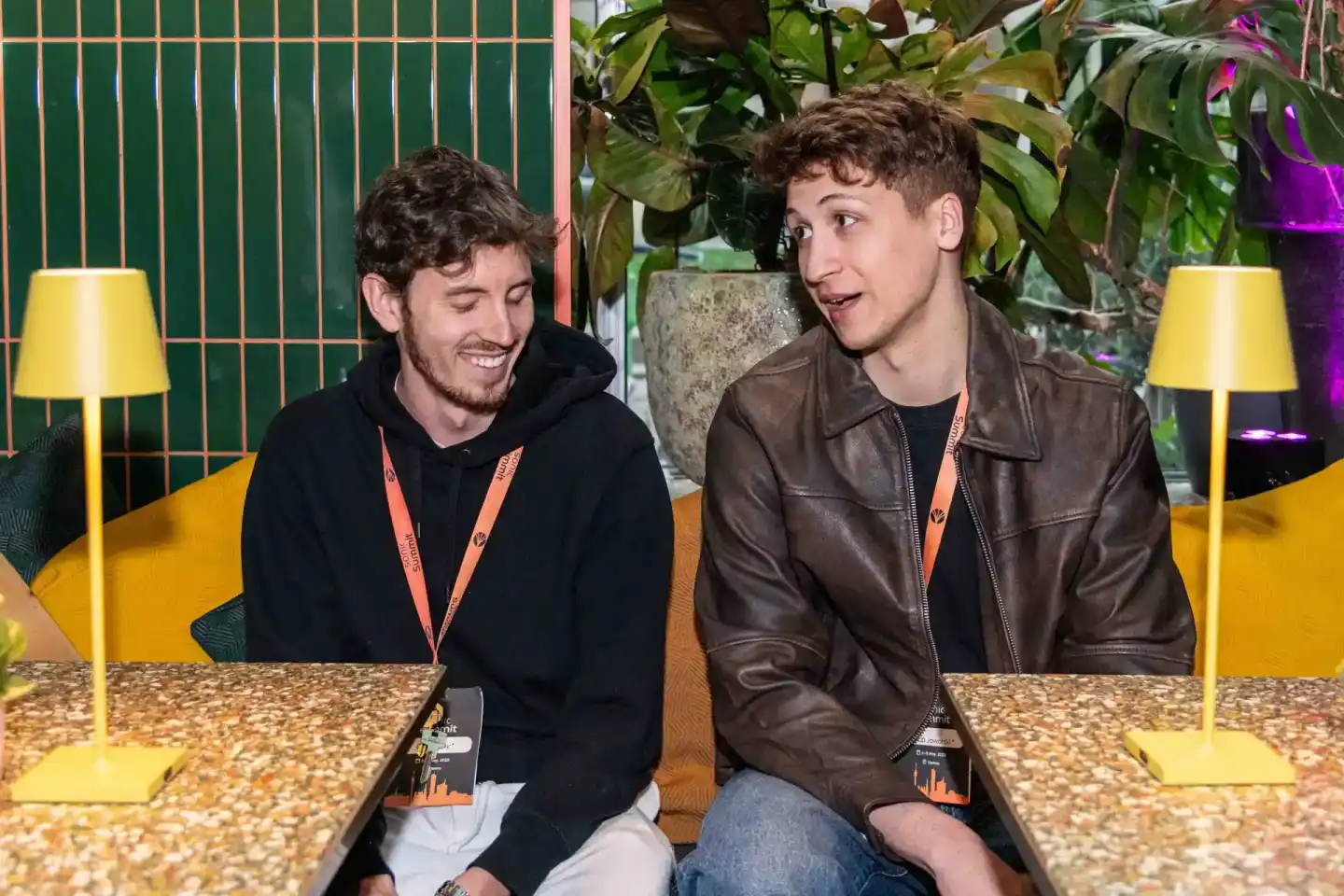 Zwei junge Männer mit Event-Badges sitzen an einem Tisch beim Sonic Summit in Wien, lachen und unterhalten sich